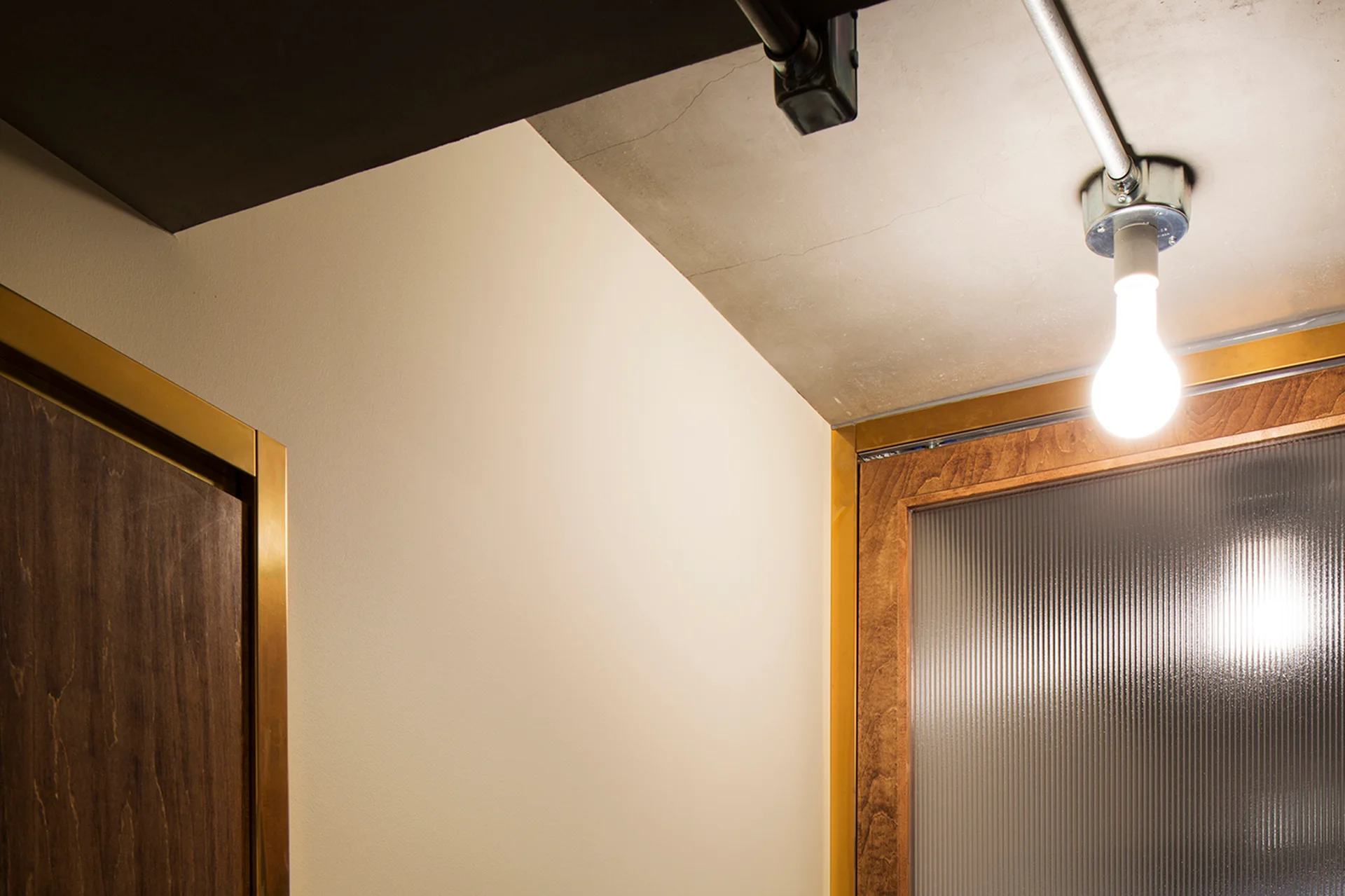 A close-up view of a concrete ceiling with an exposed light bulb and conduit next to wooden doors framed in brass trim.