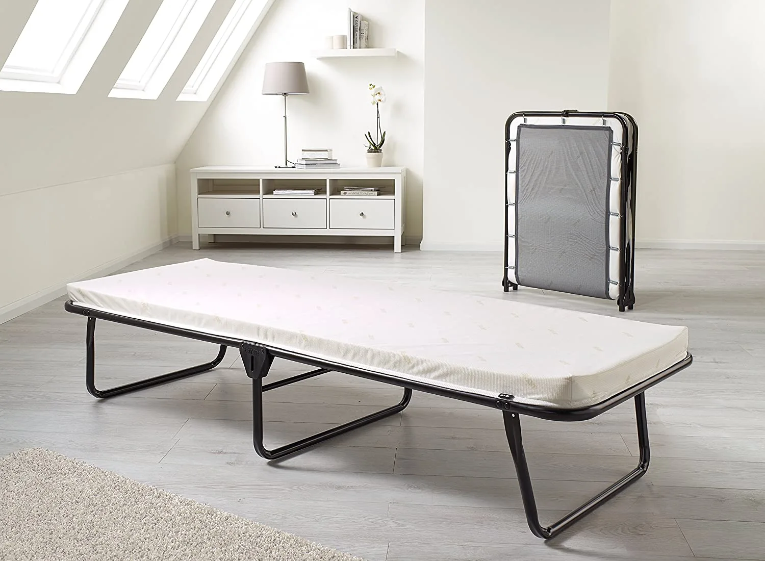 A black metal frame folding bed with a white mattress is set up in a minimalist loft bedroom with sloped skylights and grey wood flooring.
