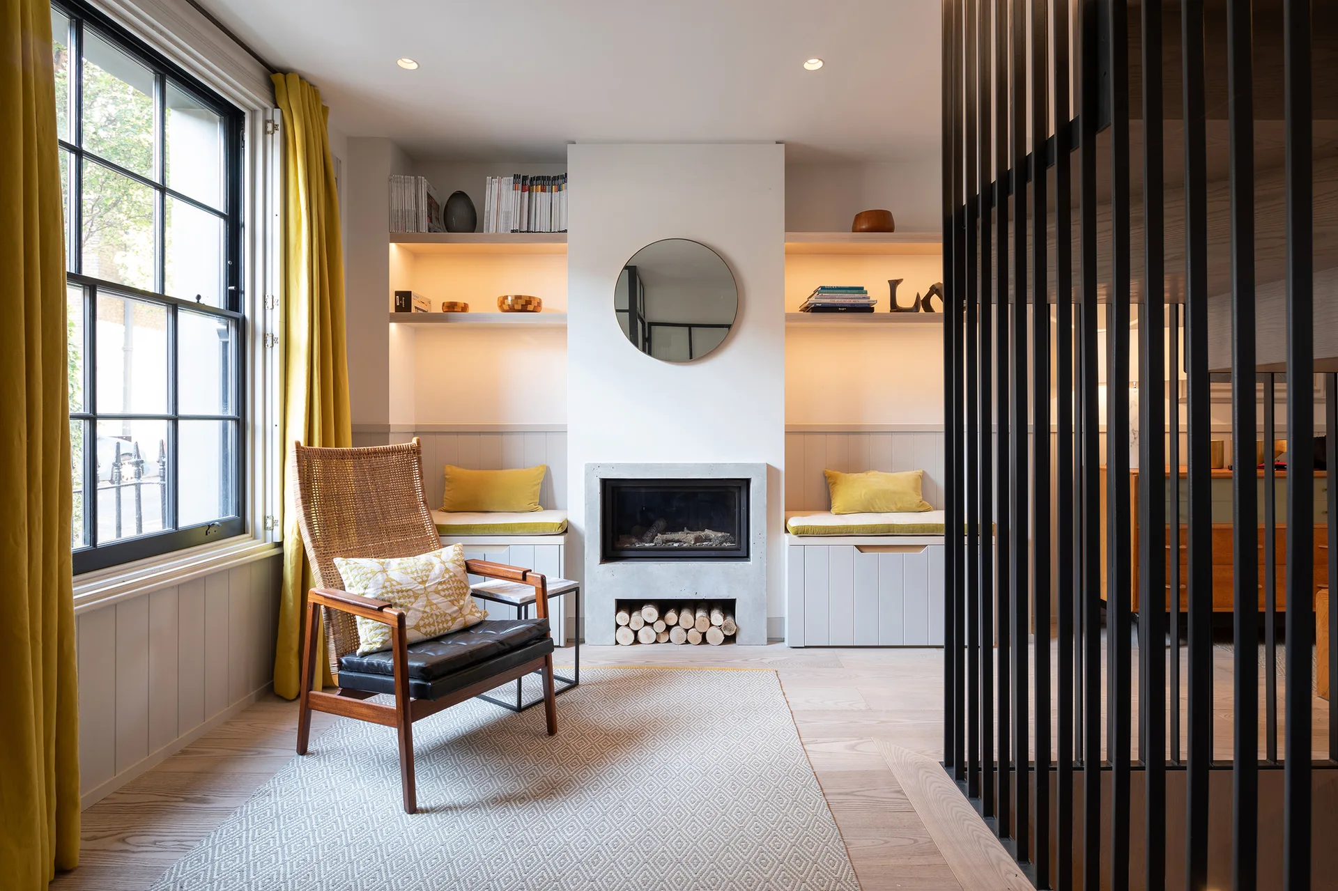 A living room features a central grey concrete fireplace with built-in bench seating, a wood and rattan armchair, and yellow curtains by a window.