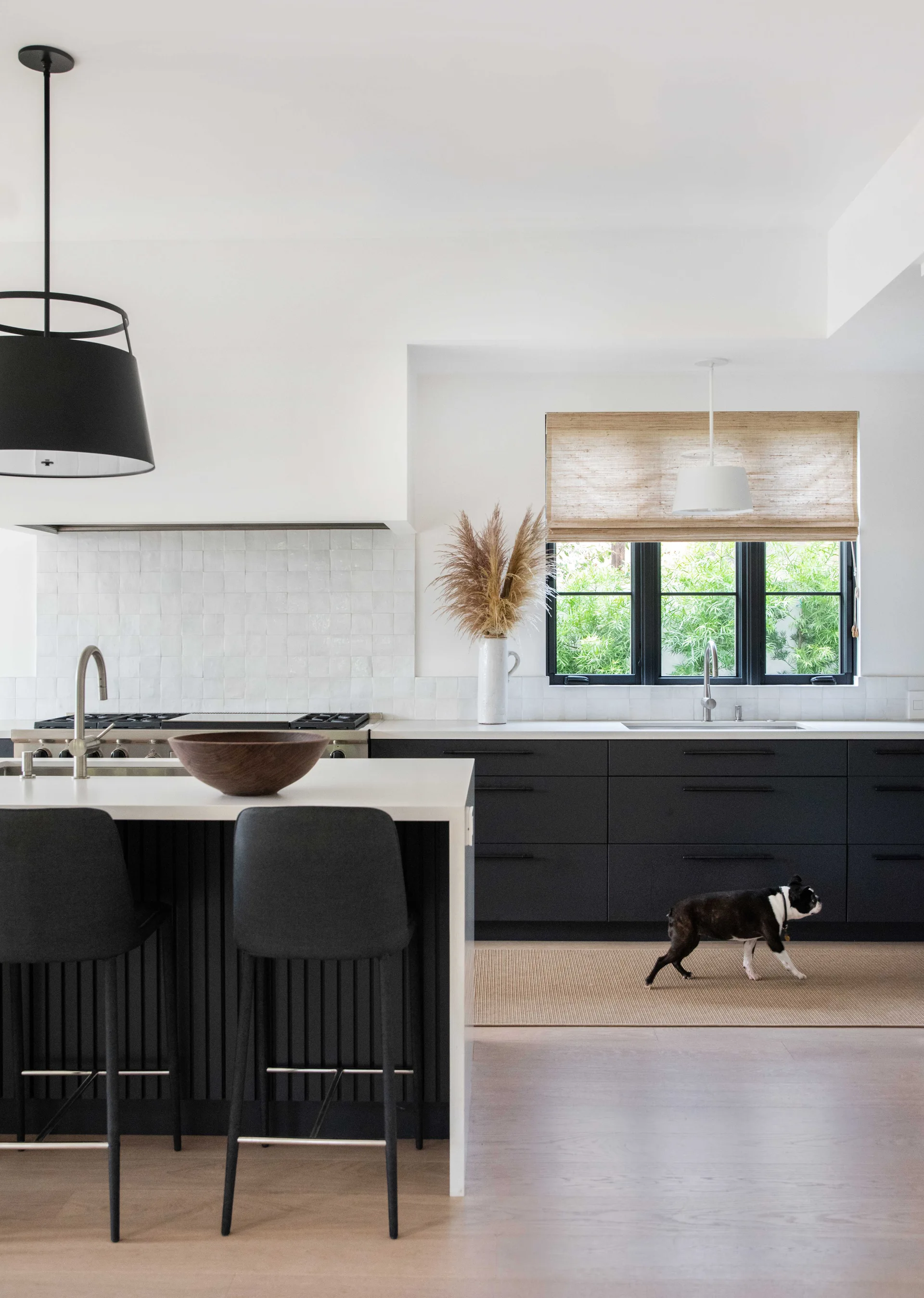 A modern kitchen featuring matte black lower cabinets, a central island with bar stools, white tiled walls, and a window with a woven shade.