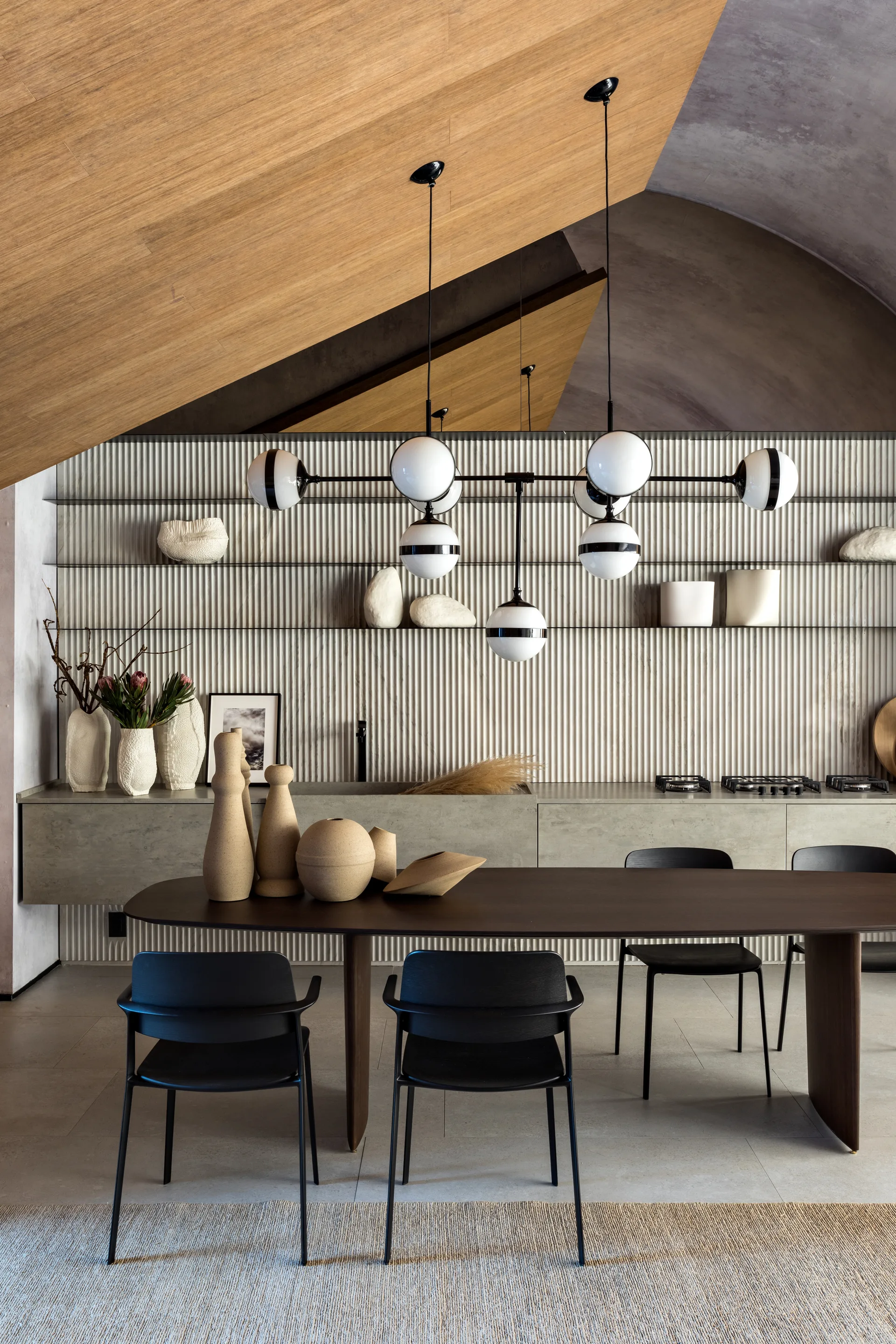 A dark wood oval dining table with black chairs sits before a light-colored fluted wall under an angled wood-paneled ceiling.