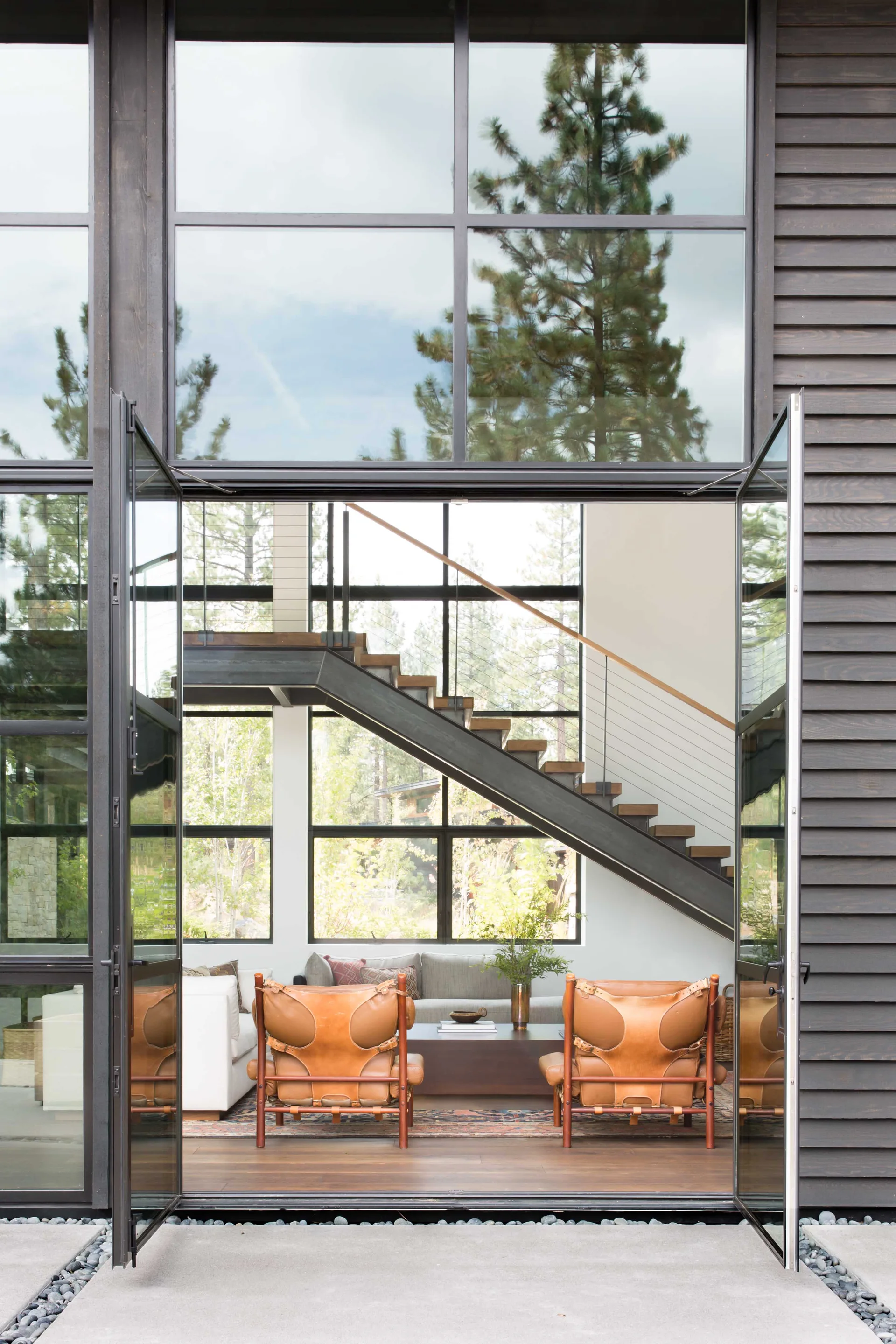 Open glass double doors look into a modern living room featuring two tan leather lounge chairs, a low coffee table, and a steel floating staircase.