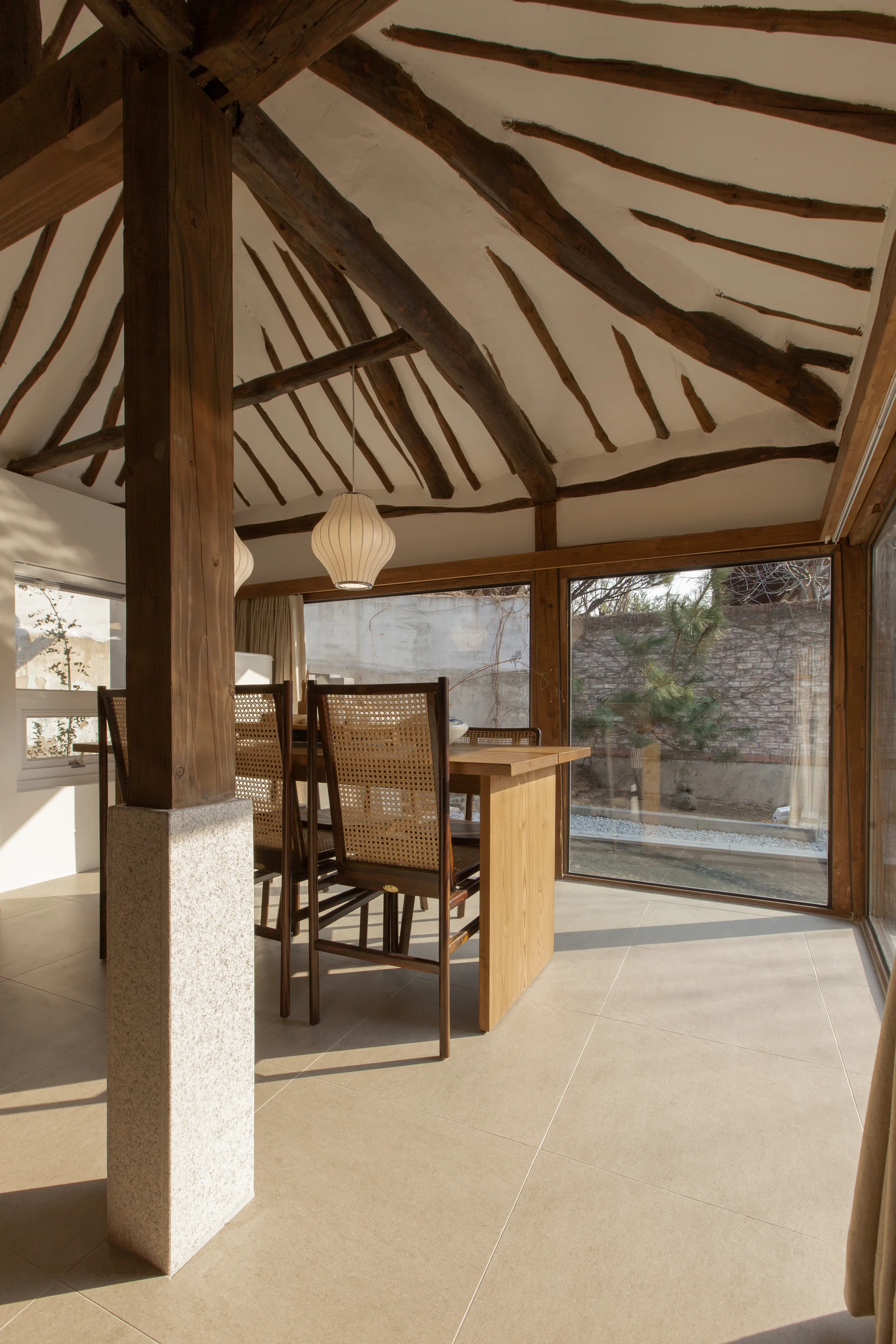 A dining room features a wooden table and caned chairs beneath a vaulted ceiling with radiating rustic timber beams next to large windows.