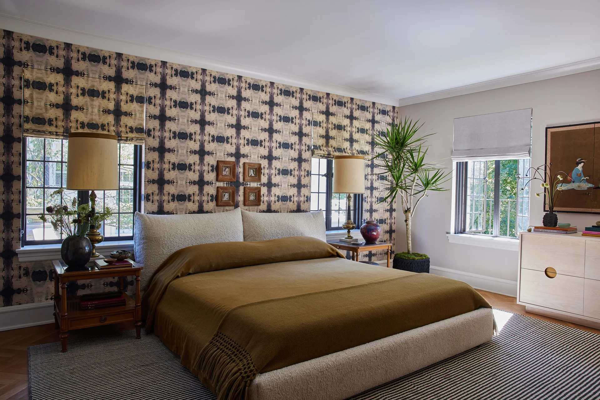A bedroom features an upholstered boucle bed with a brown coverlet against an abstract patterned wallpaper accent wall and a striped area rug.