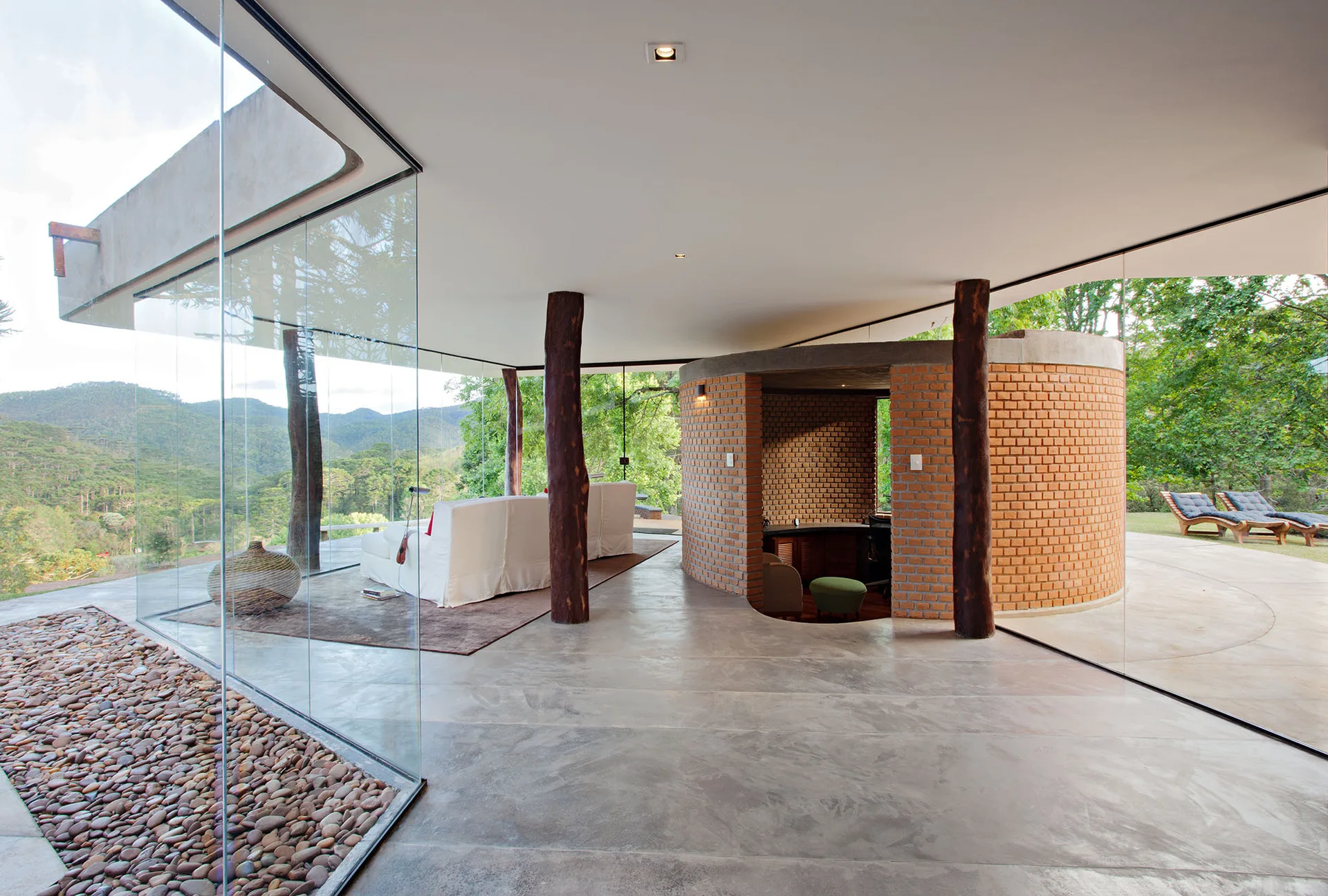 A modern open living room with polished concrete flooring, floor-to-ceiling glass walls, natural wood columns, and a curved red brick partition.