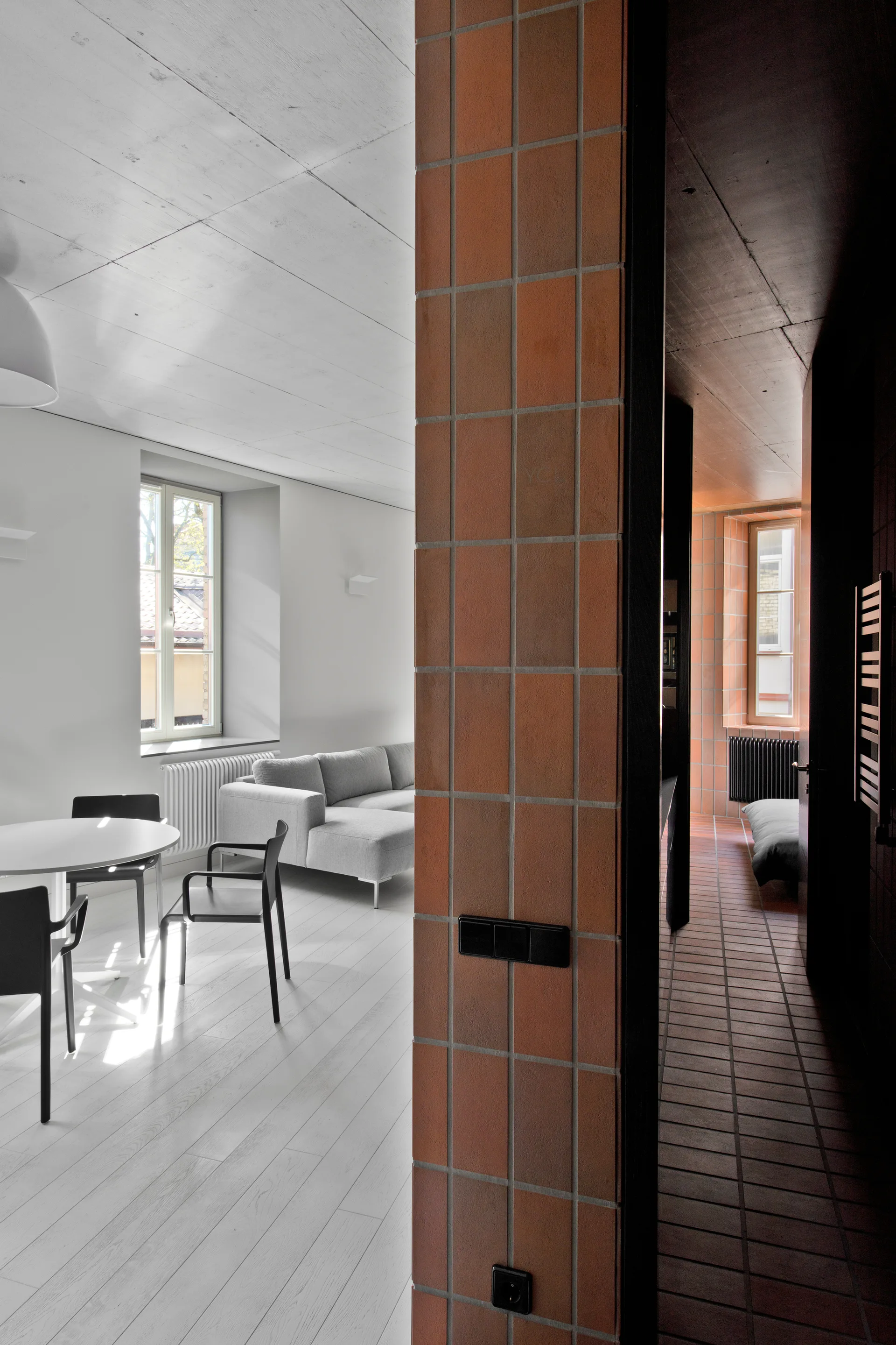 A minimalist living room with a grey sofa is separated from a dark corridor by a red tiled partition wall.