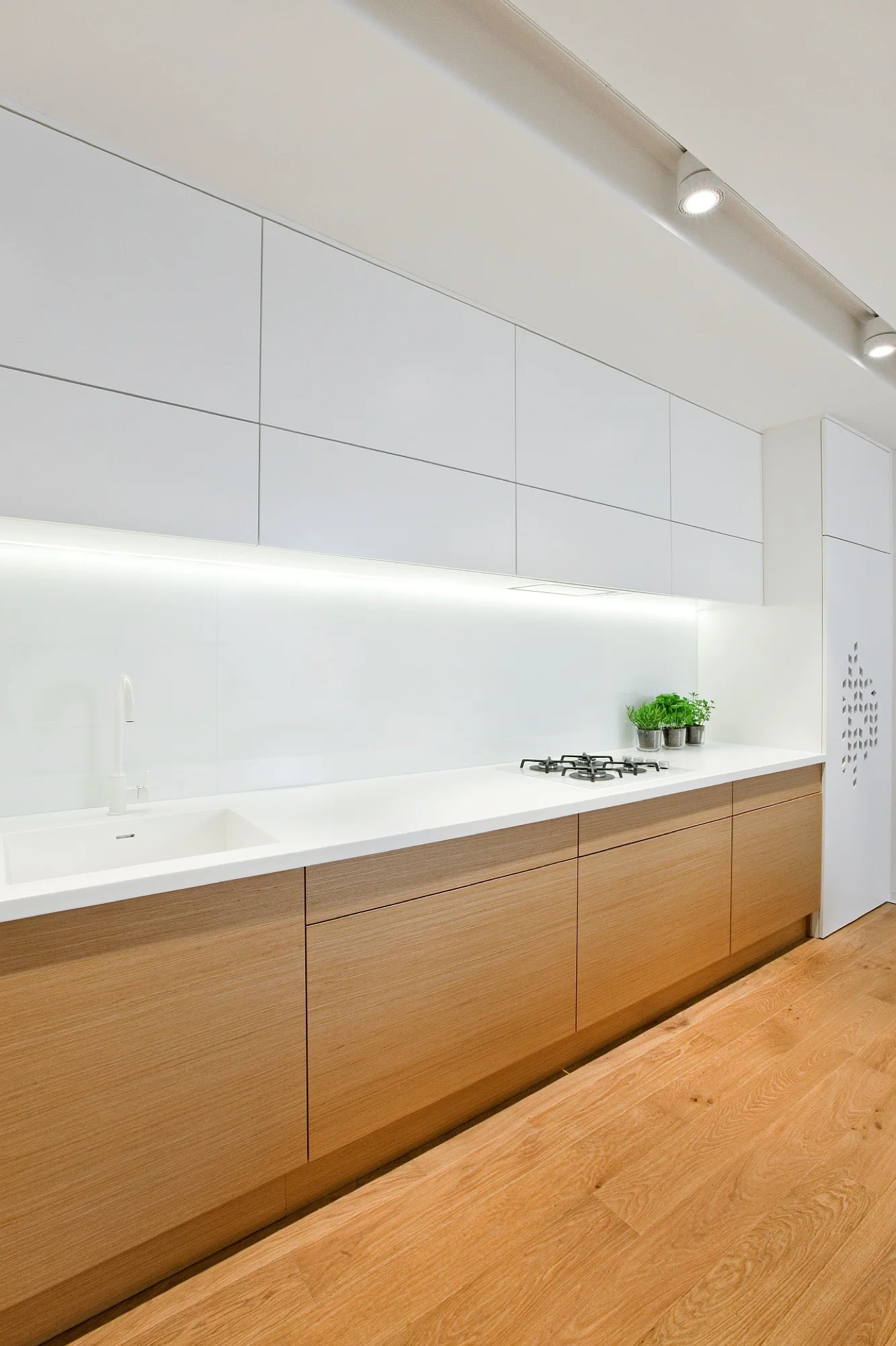 A modern kitchen features white upper cabinets, flat-panel wood base drawers, a white countertop with an integrated sink, and under-cabinet LED lighting.