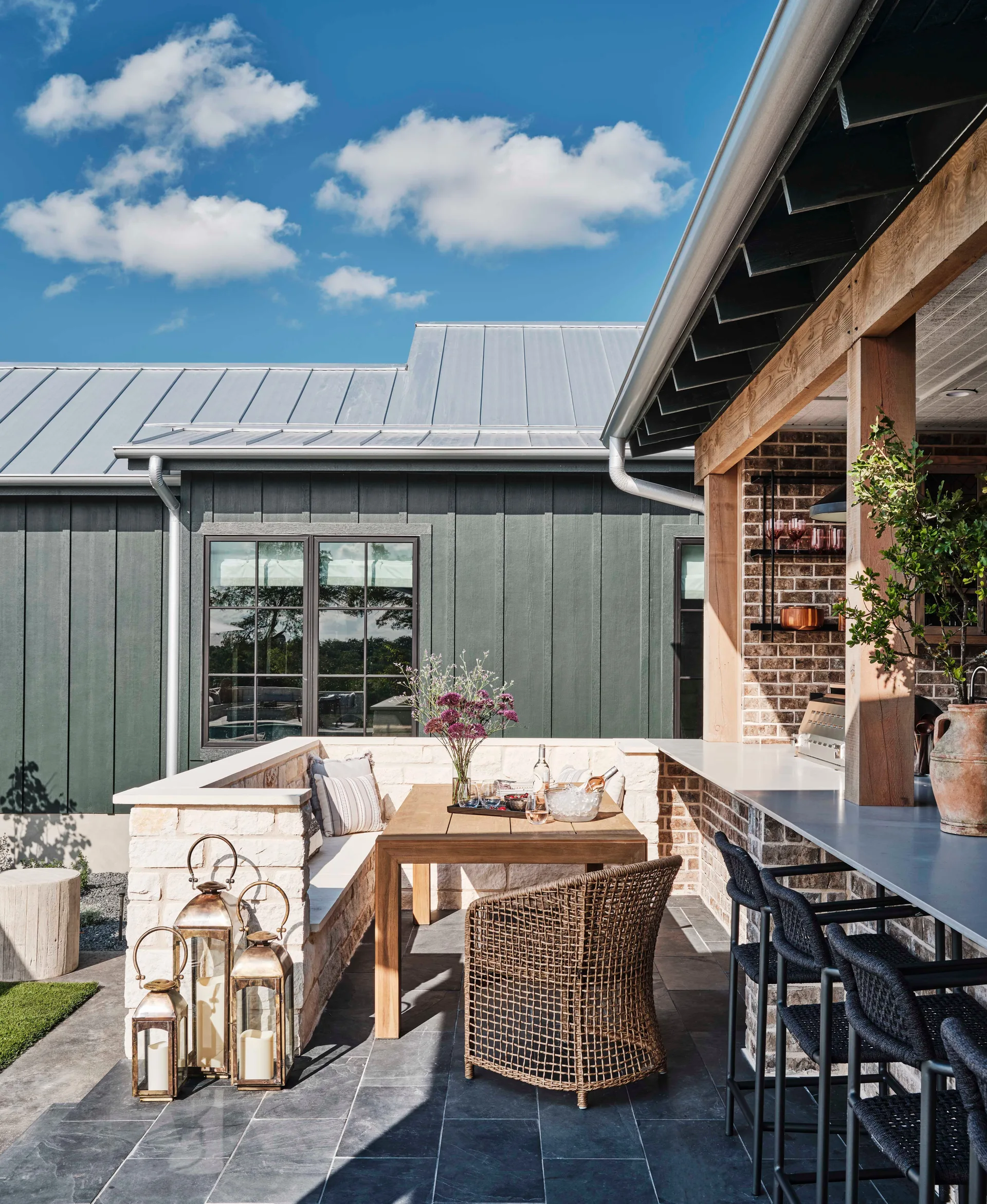 An outdoor dining patio features a wooden table with built-in stone bench seating, a covered bar with wicker stools, and dark siding.