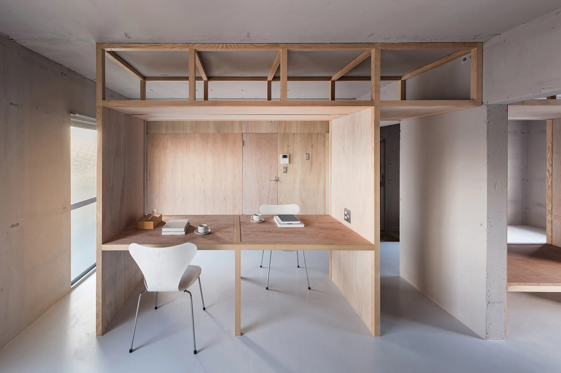 A minimalist home office featuring a built-in plywood desk structure with a wooden overhead grid, grey concrete walls, and two white chairs.