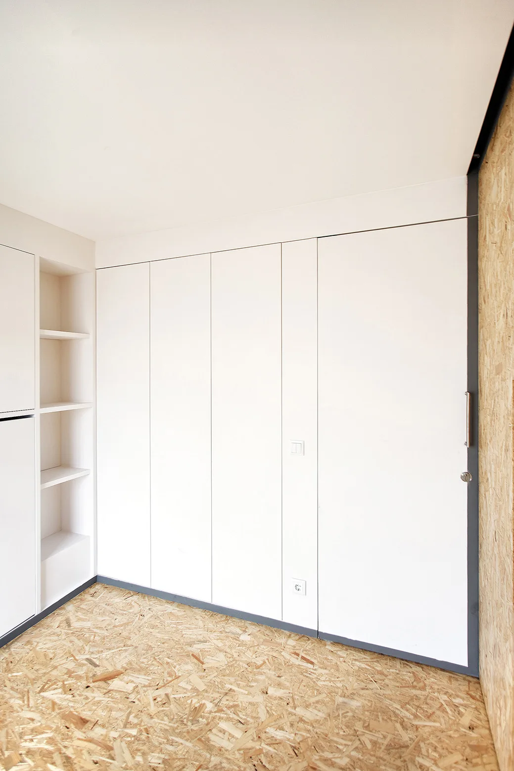 A wall of seamless white built-in cabinetry stands next to a recessed shelving niche above oriented strand board flooring.