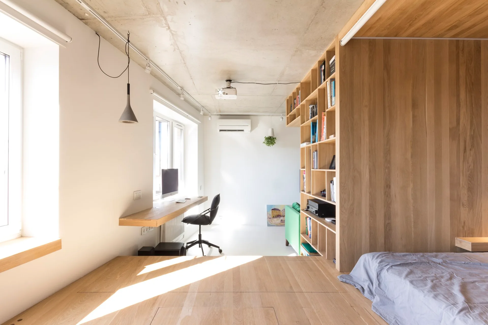 A modern studio bedroom featuring a raised light wood platform bed, an integrated wooden bookcase partition, and a floating desk.