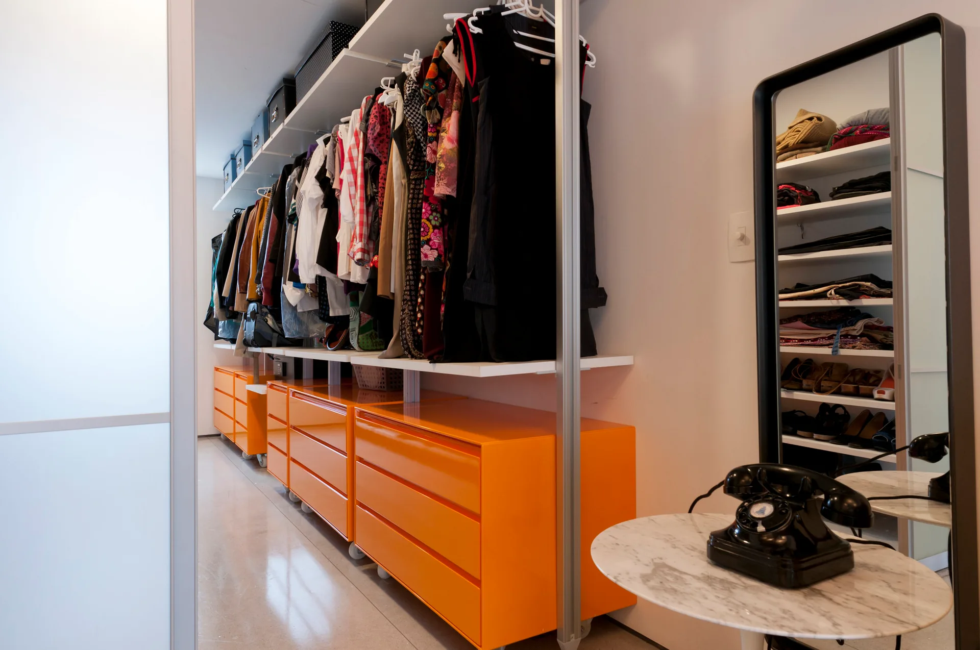 A walk-in closet with glossy orange mobile drawers beneath hanging clothes, a black-framed mirror, and a round marble side table.