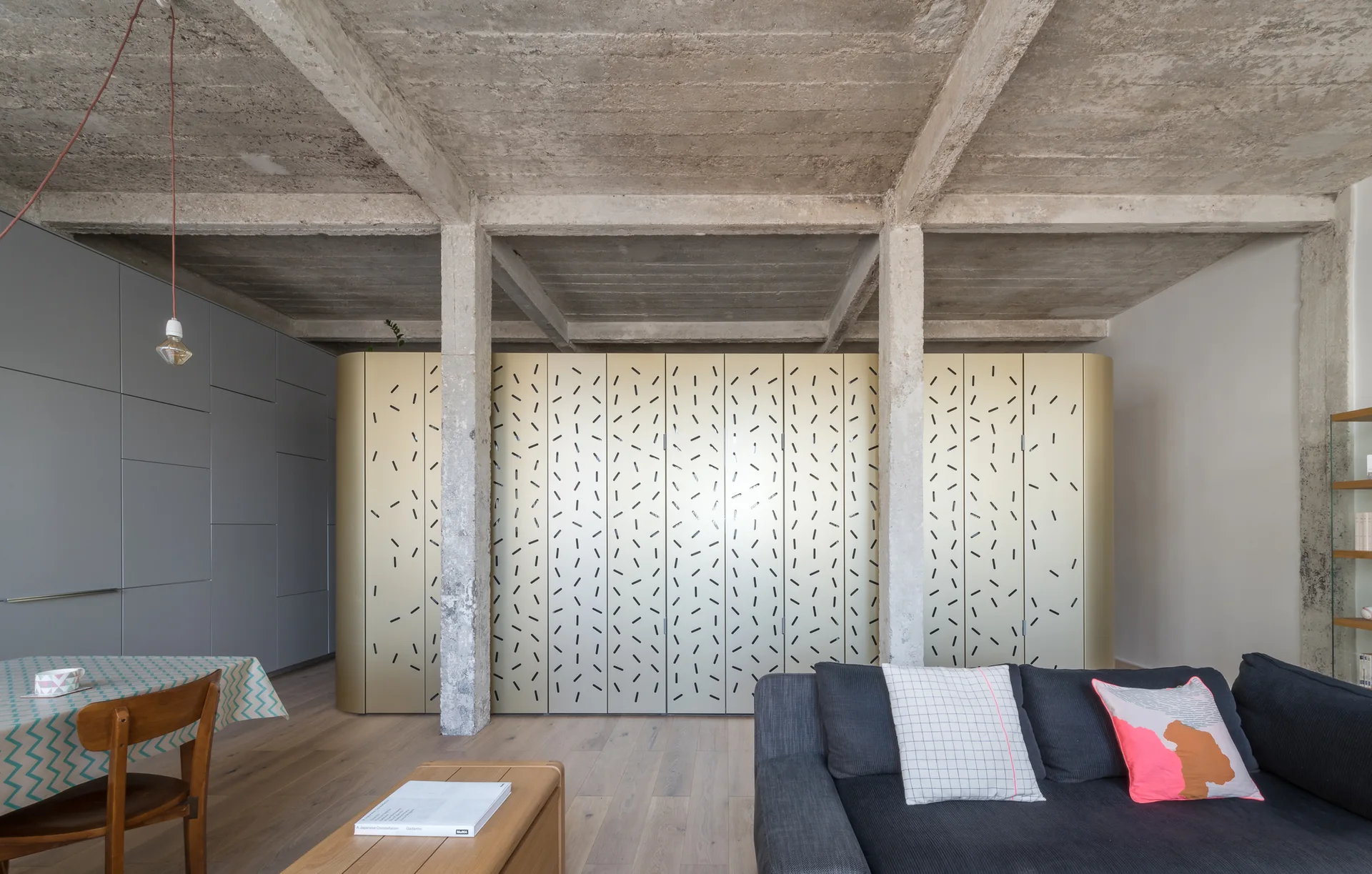 An open-plan living room features a dark grey sofa, a gold patterned cabinet wall, and an exposed concrete ceiling with structural columns.