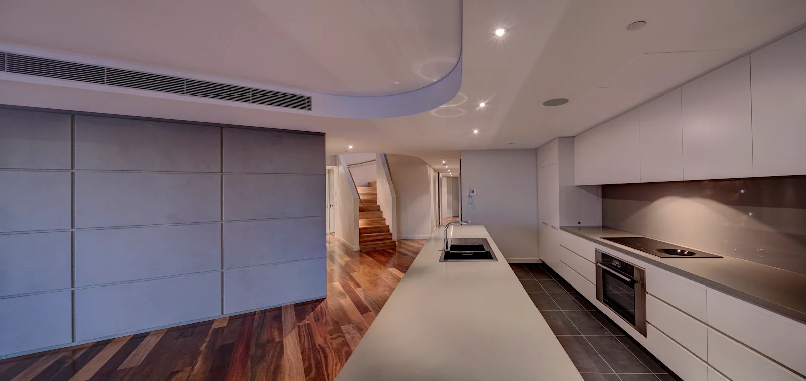 A minimalist kitchen features white flat-front cabinets, a long island with a black sink, a curved ceiling detail, and dark hardwood floors.