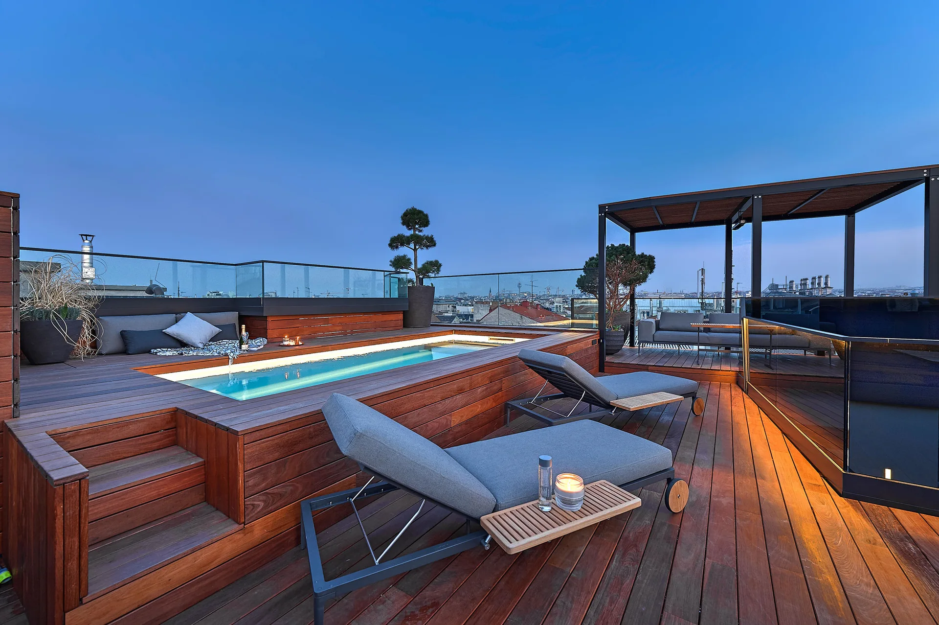 A rooftop terrace at dusk featuring a wooden deck with an integrated swimming pool, gray chaise lounges, and glass railings overlooking a city skyline.