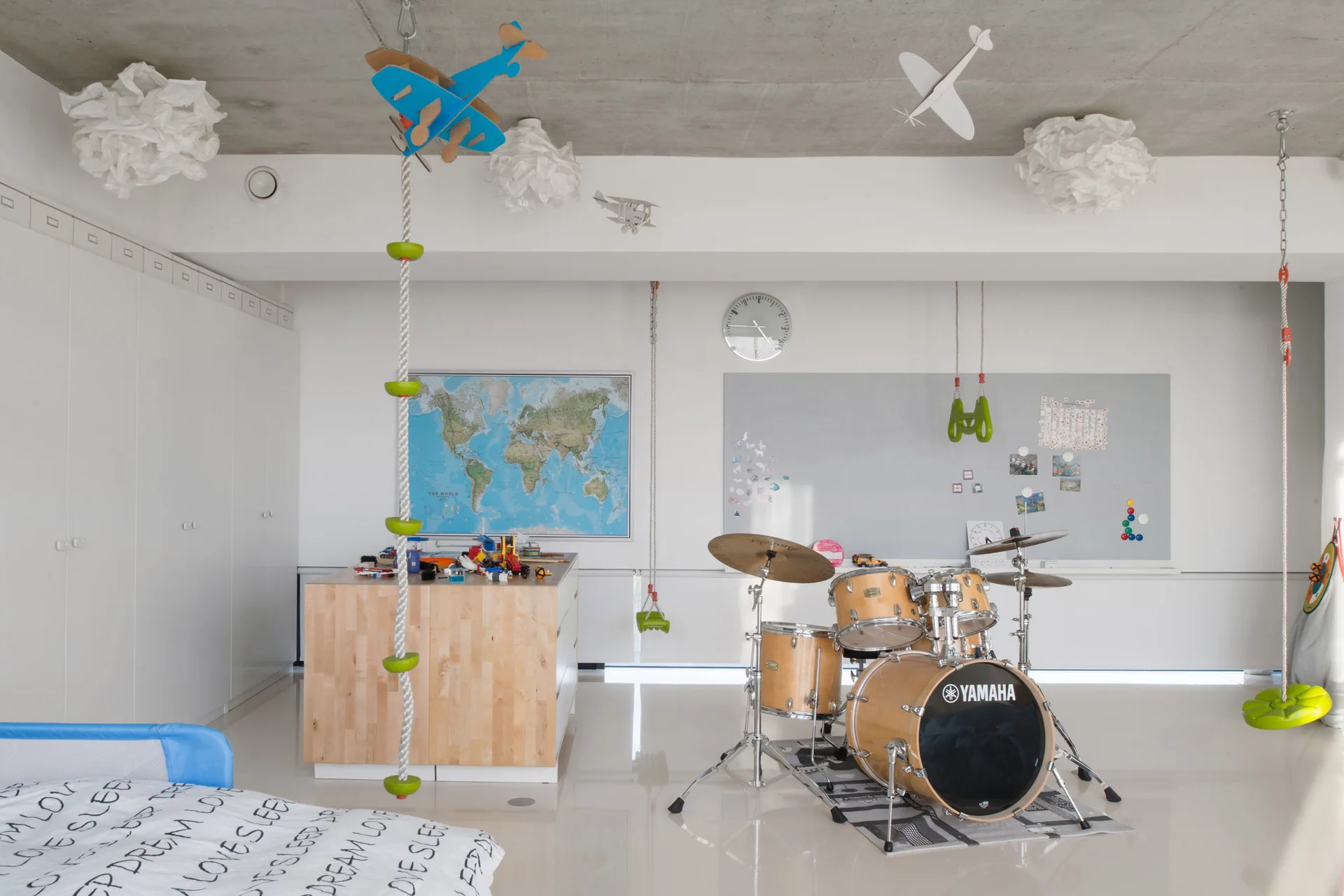 A modern kids playroom featuring a drum set on a graphic rug, ceiling-mounted green play equipment, and a wooden craft table under a concrete ceiling.