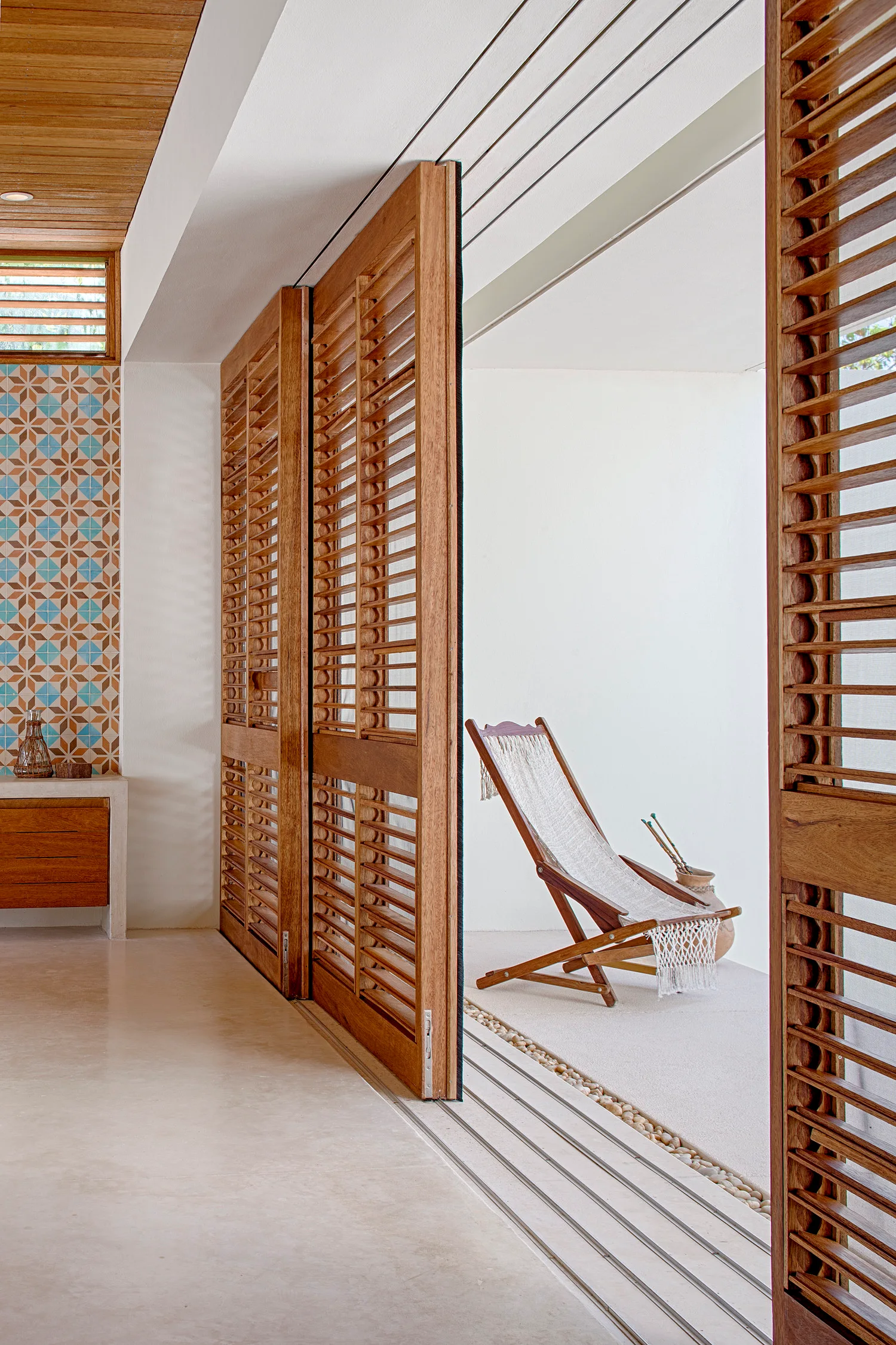 Warm wooden louvered sliding doors open from a light concrete room to a white-walled patio with a woven deck chair.