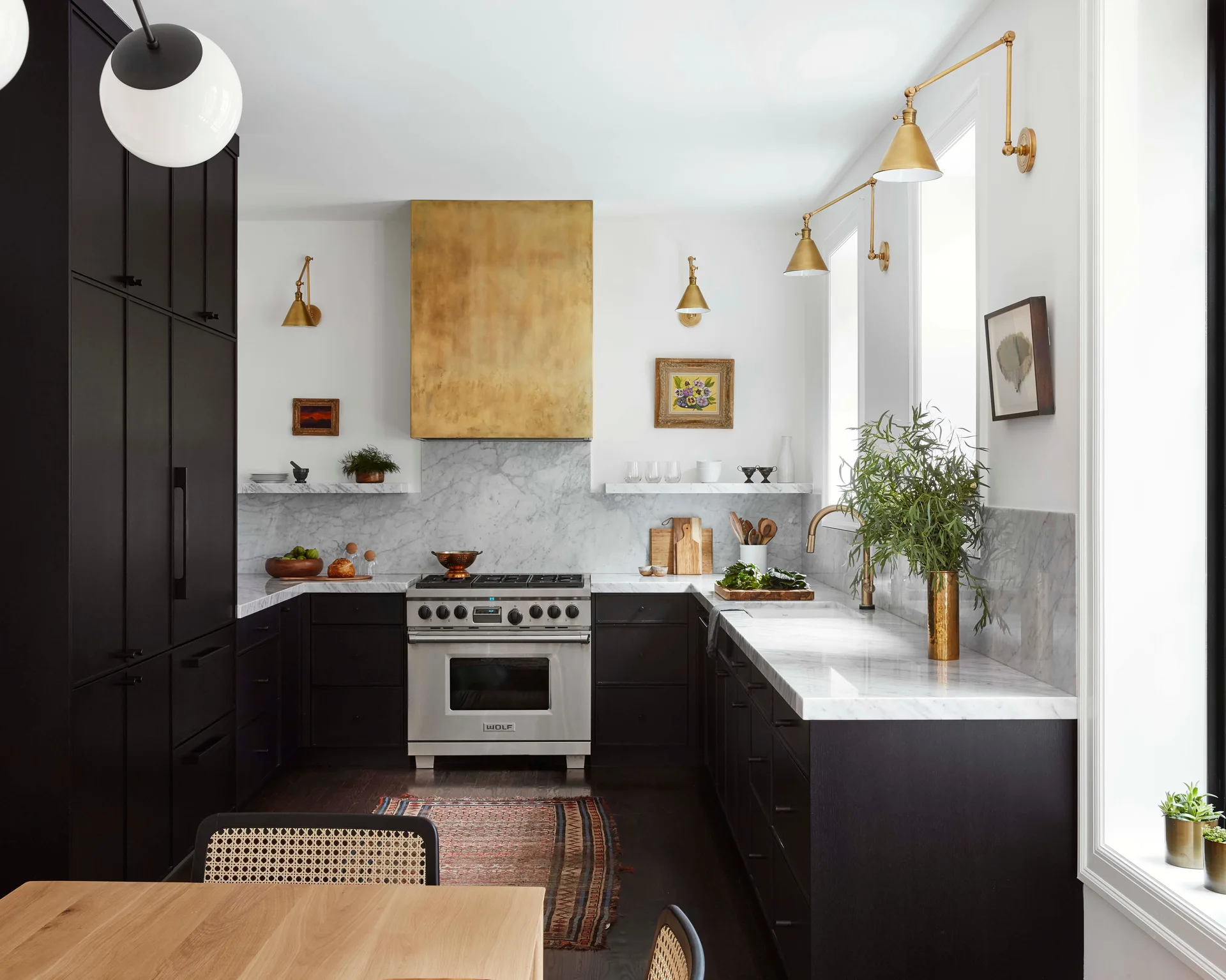 A kitchen features matte black cabinets, a custom brass range hood over a stainless steel stove, and white marble countertops with a matching backsplash.