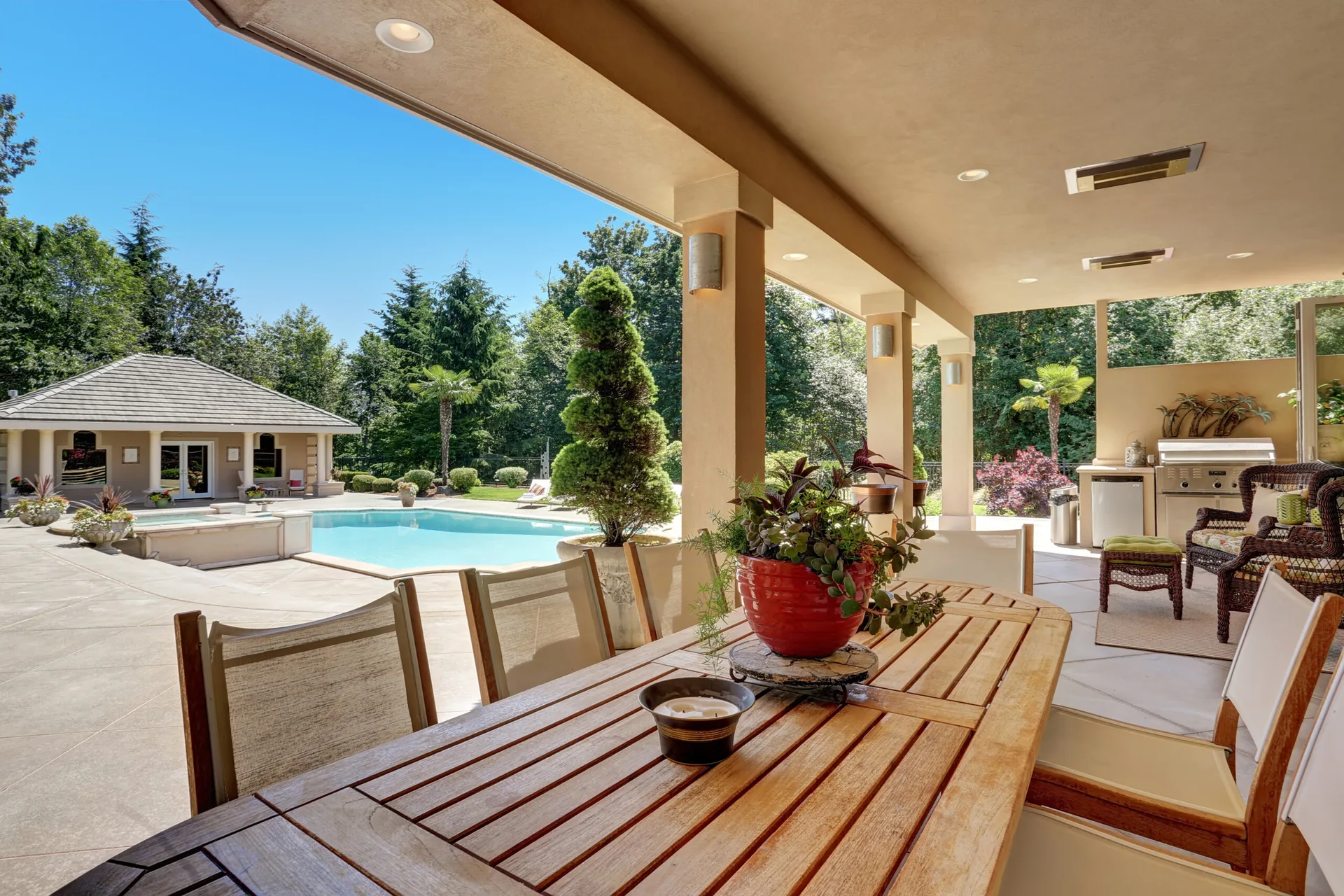 A covered outdoor patio features a slatted wood dining table with chairs, beige stucco pillars, and a swimming pool in the background.