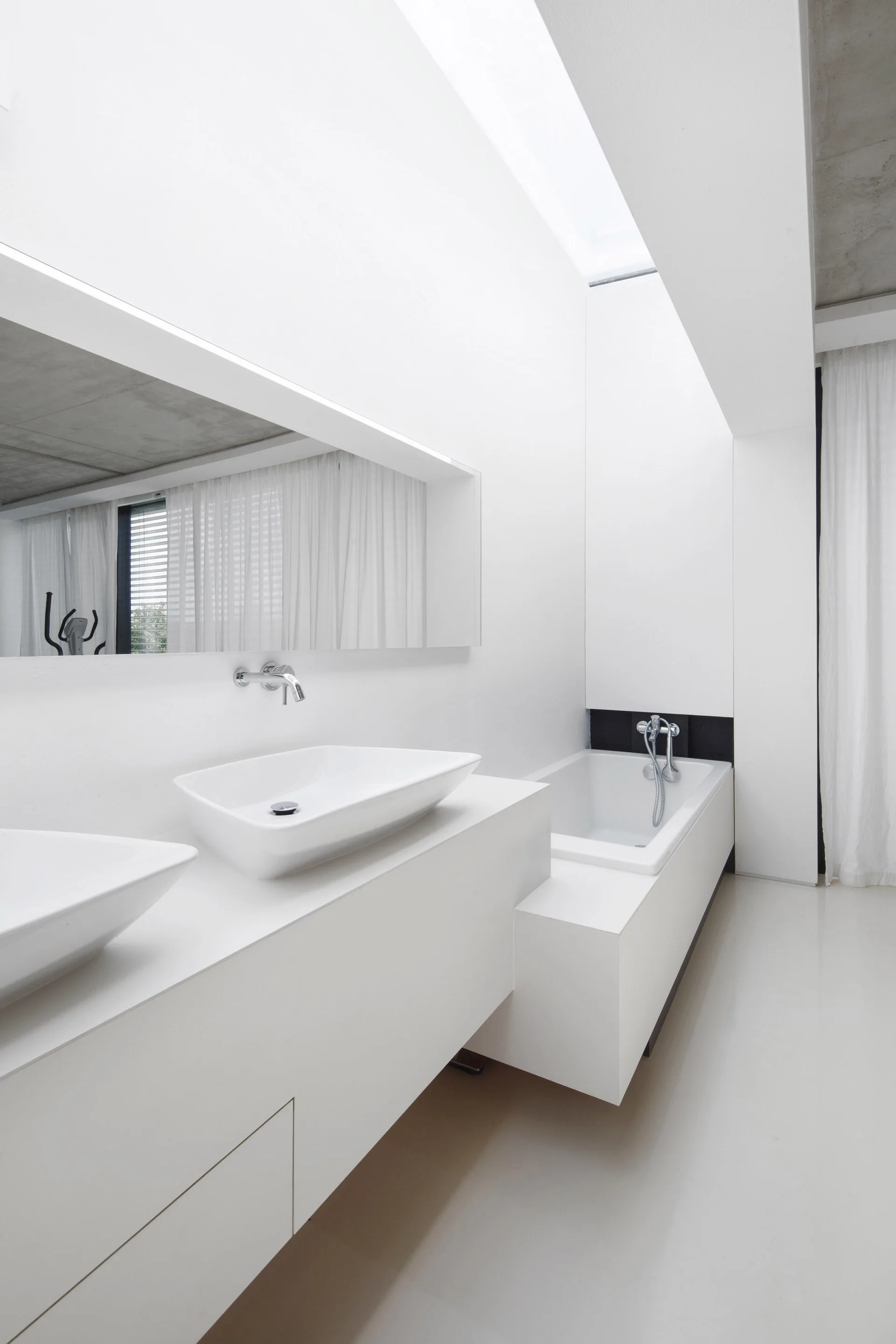 A modern white bathroom features two ceramic vessel sinks on a long built-in vanity and a bathtub beneath an overhead skylight.