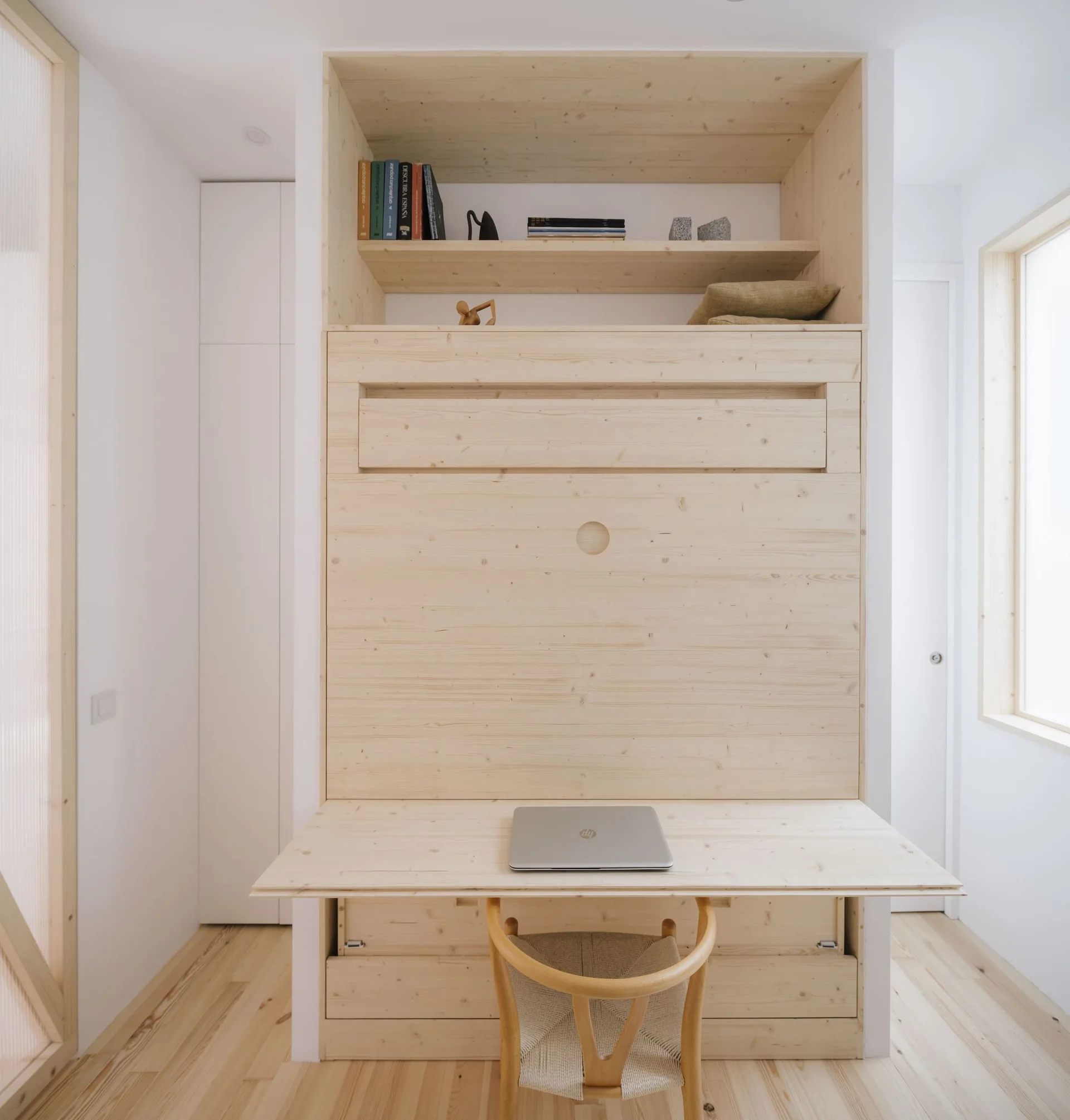 A minimalist home office featuring a tall light wood built-in unit with an integrated desk, open shelving, and a wooden chair on matching plank floors.