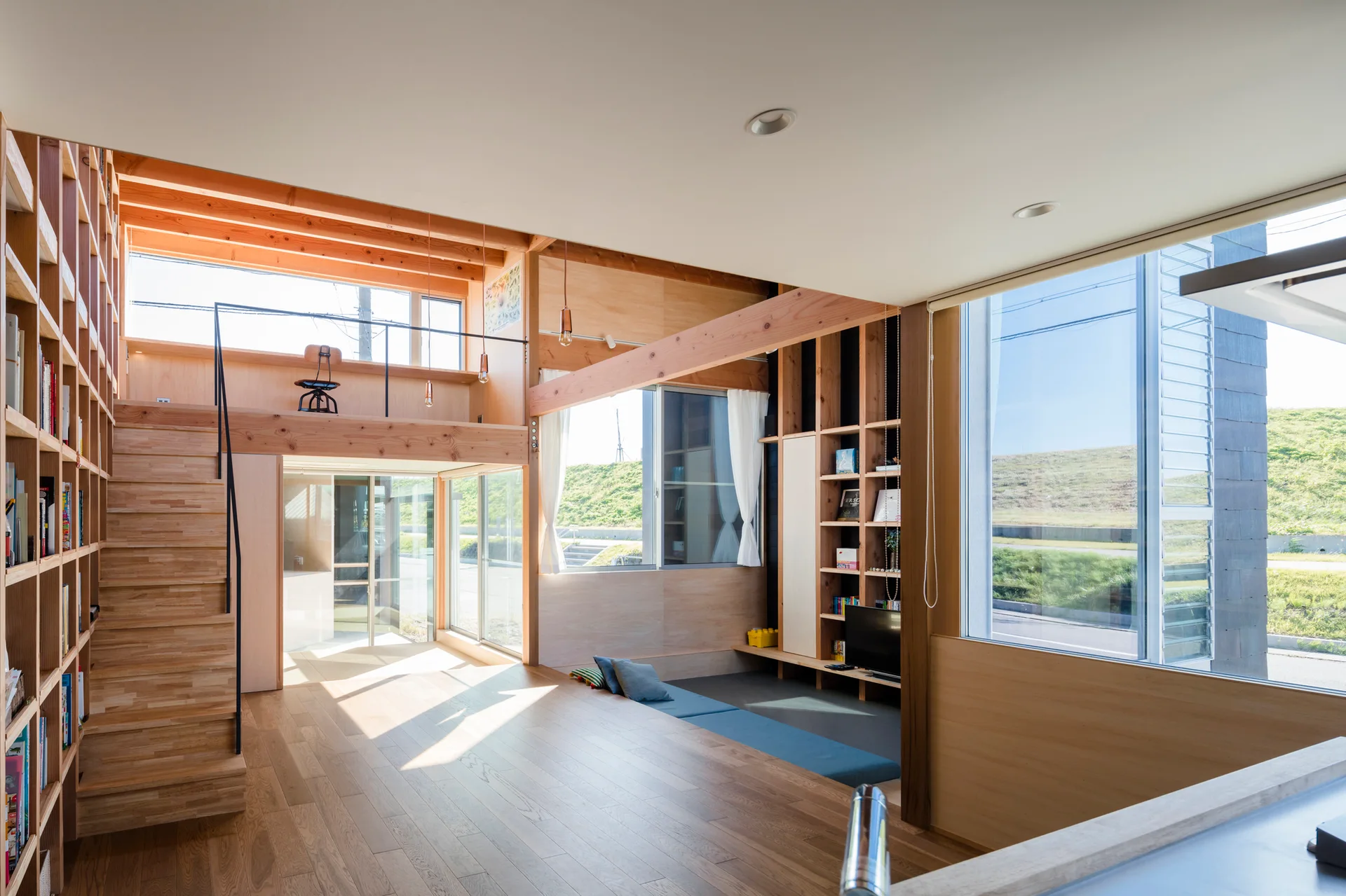 A bright double-height living room with natural wood wall paneling, built-in bookshelves, a wooden staircase, and a mezzanine loft.