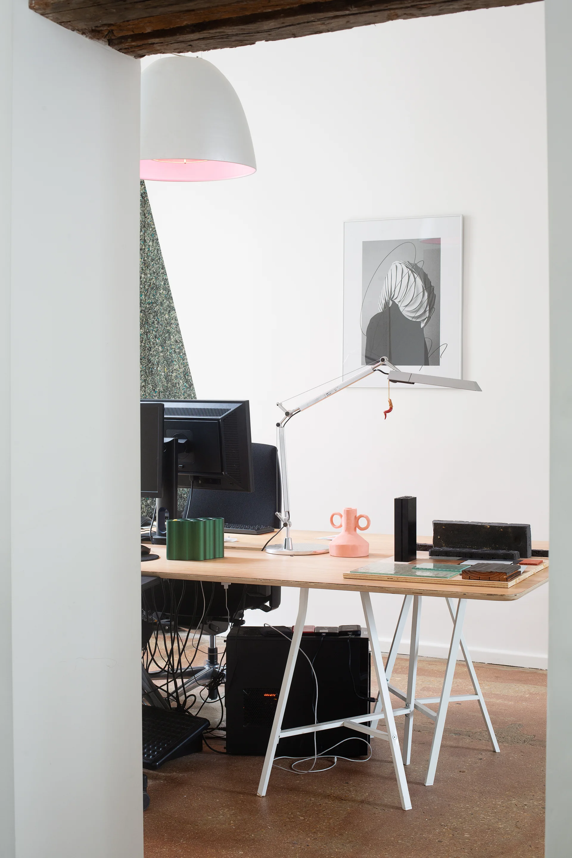 A minimalist home office features a plywood-edged trestle desk with dual monitors, a pink vase, and a task lamp against white walls.