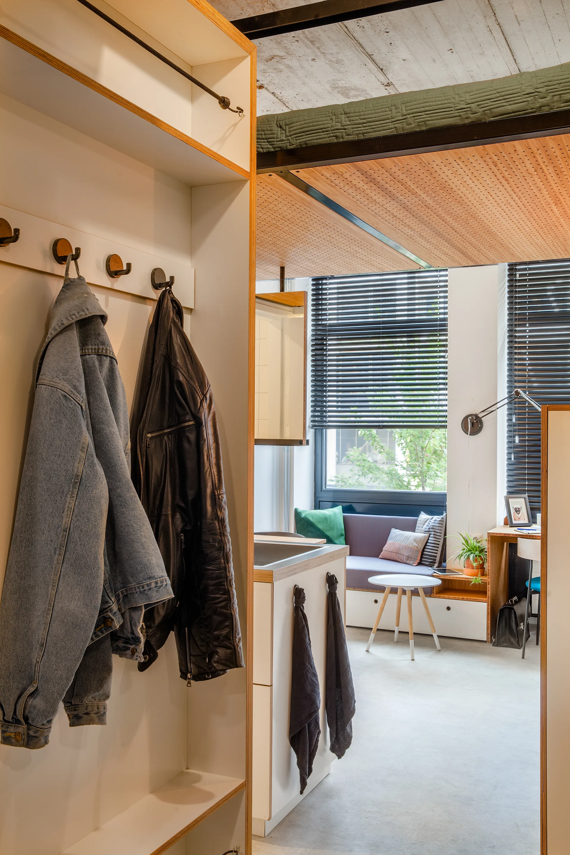 A modern studio apartment featuring white entryway storage with coat hooks, a compact kitchenette, and a lounge area under a concrete ceiling.