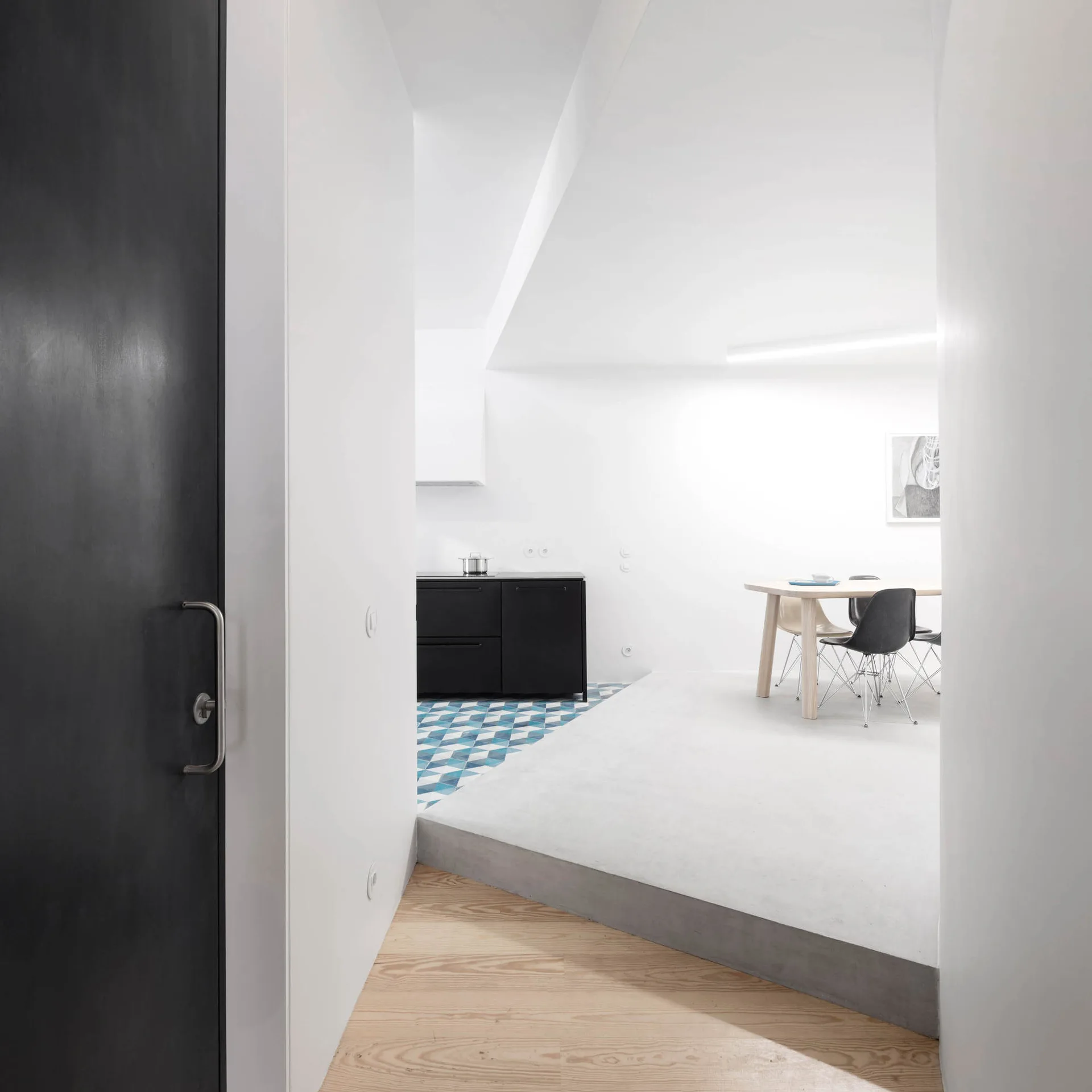 A split-level interior with wood flooring leading to a raised concrete kitchen platform decorated with blue geometric tiles and a dining set.