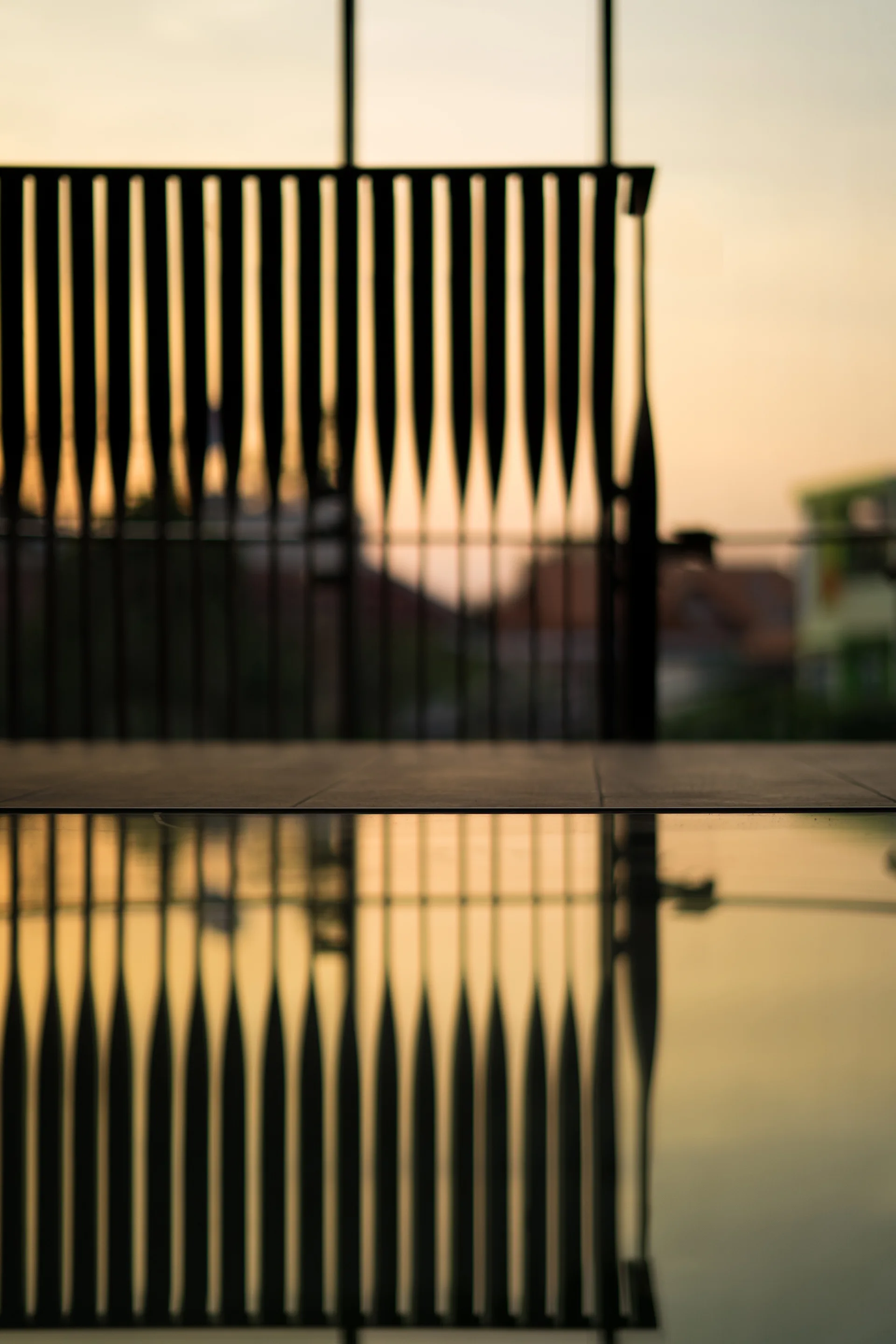 A dark slatted railing is silhouetted against a golden sunset sky, with its geometric pattern reflected perfectly in the still water of a pool.
