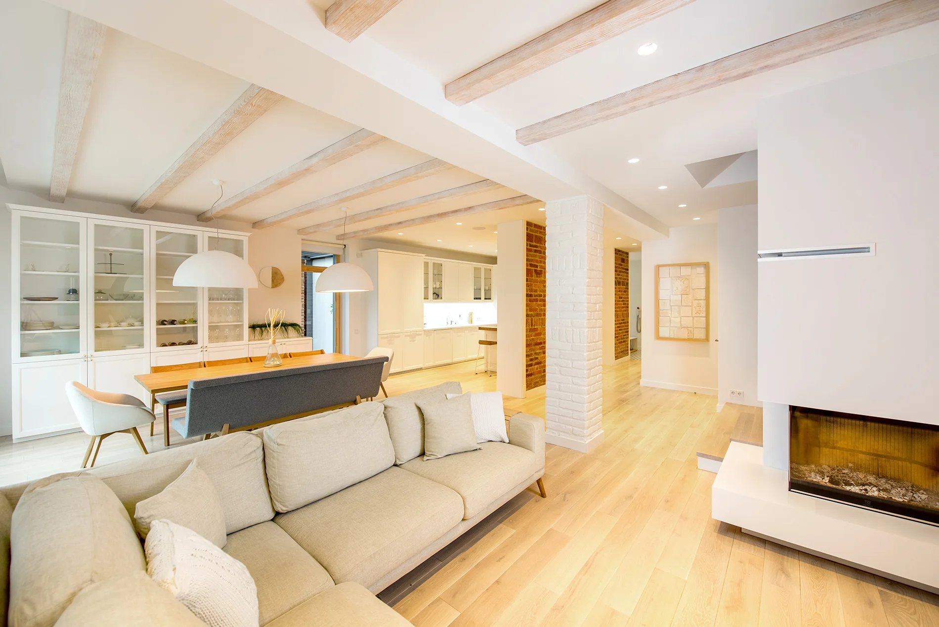 An open-plan living room features a beige sectional sofa, a dining area with pendant lights, exposed ceiling beams, and a modern white fireplace.