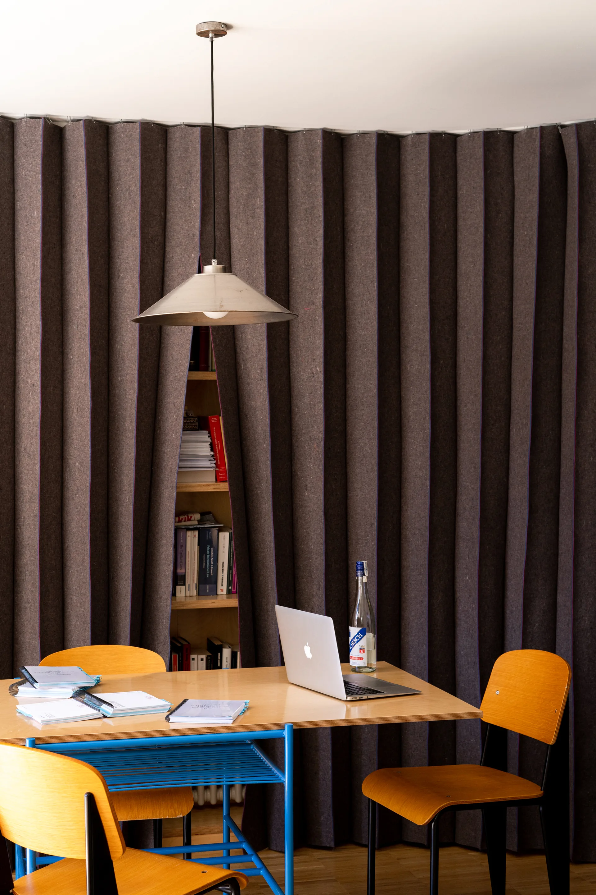 A home office features a wooden desk with a laptop and plywood chairs positioned before a floor-to-ceiling brown pleated felt curtain.
