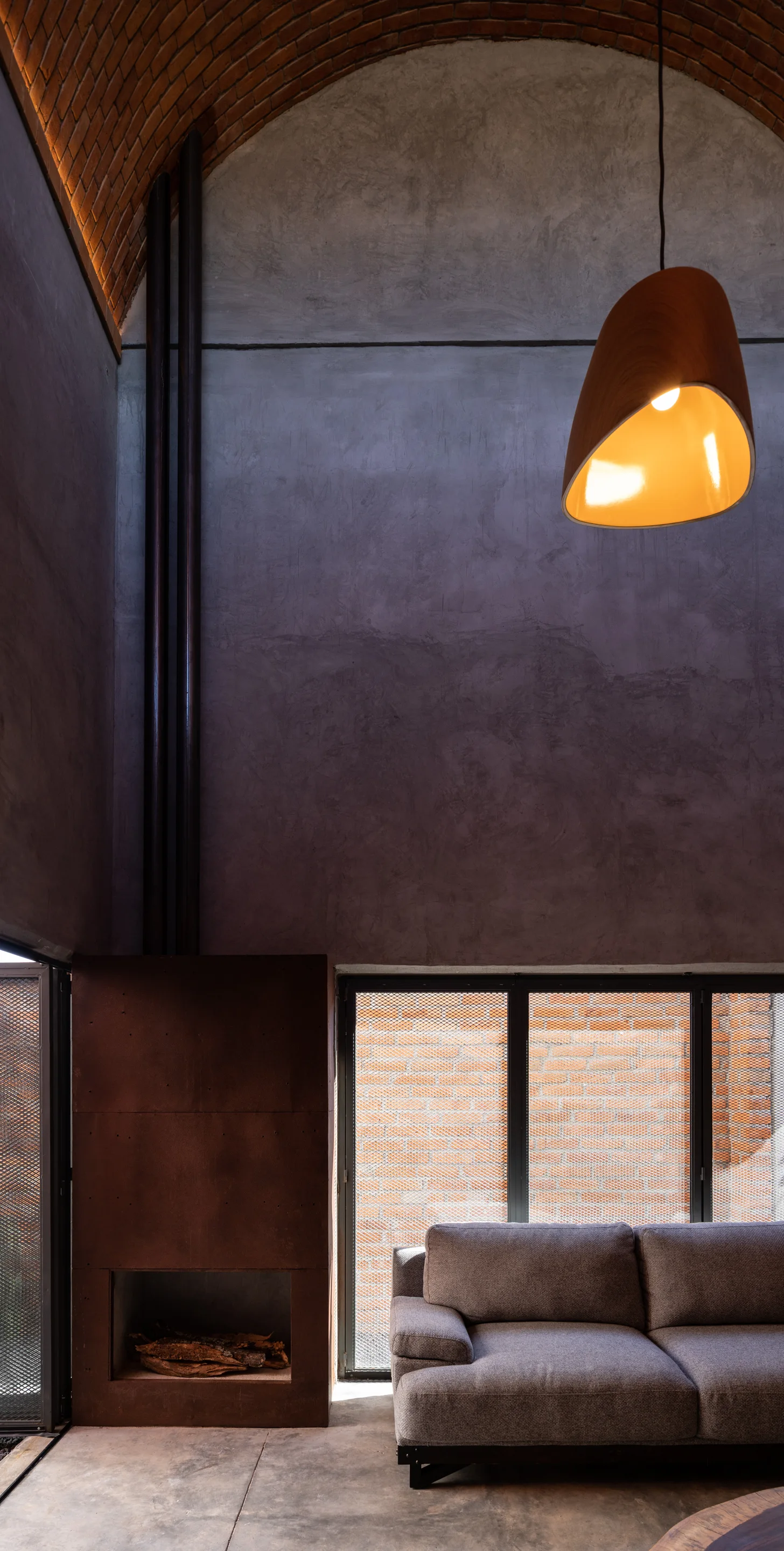 A grey sofa sits next to a rust-colored metal fireplace in an industrial living room featuring a vaulted brick ceiling and a smooth concrete wall.