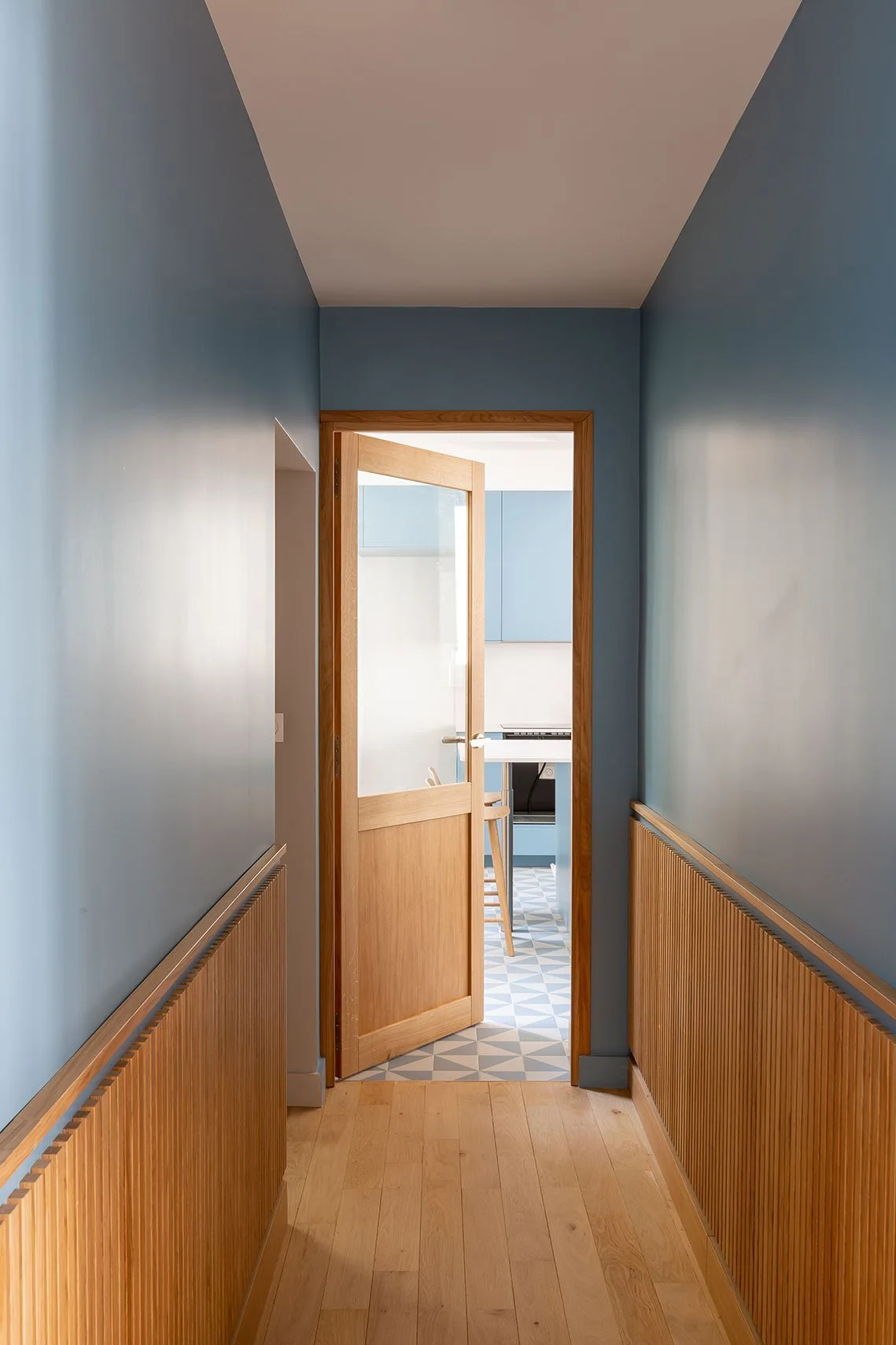 A narrow blue hallway with vertical wood slat wainscoting leads through an open glass door into a kitchen with geometric tile flooring.