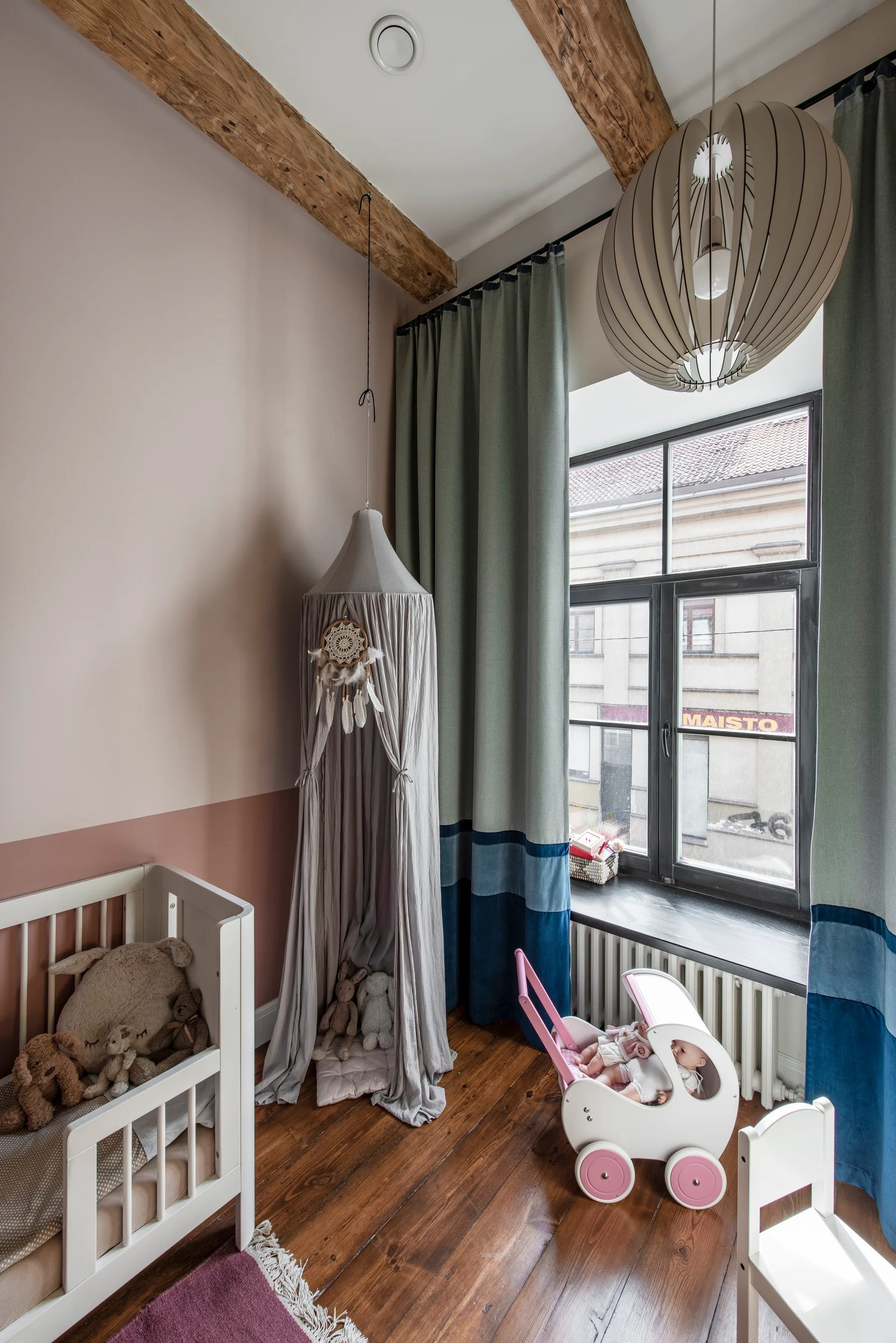A nursery features a white wooden crib, a fabric canopy hanging from a ceiling beam, and two-tone pink walls next to a window.