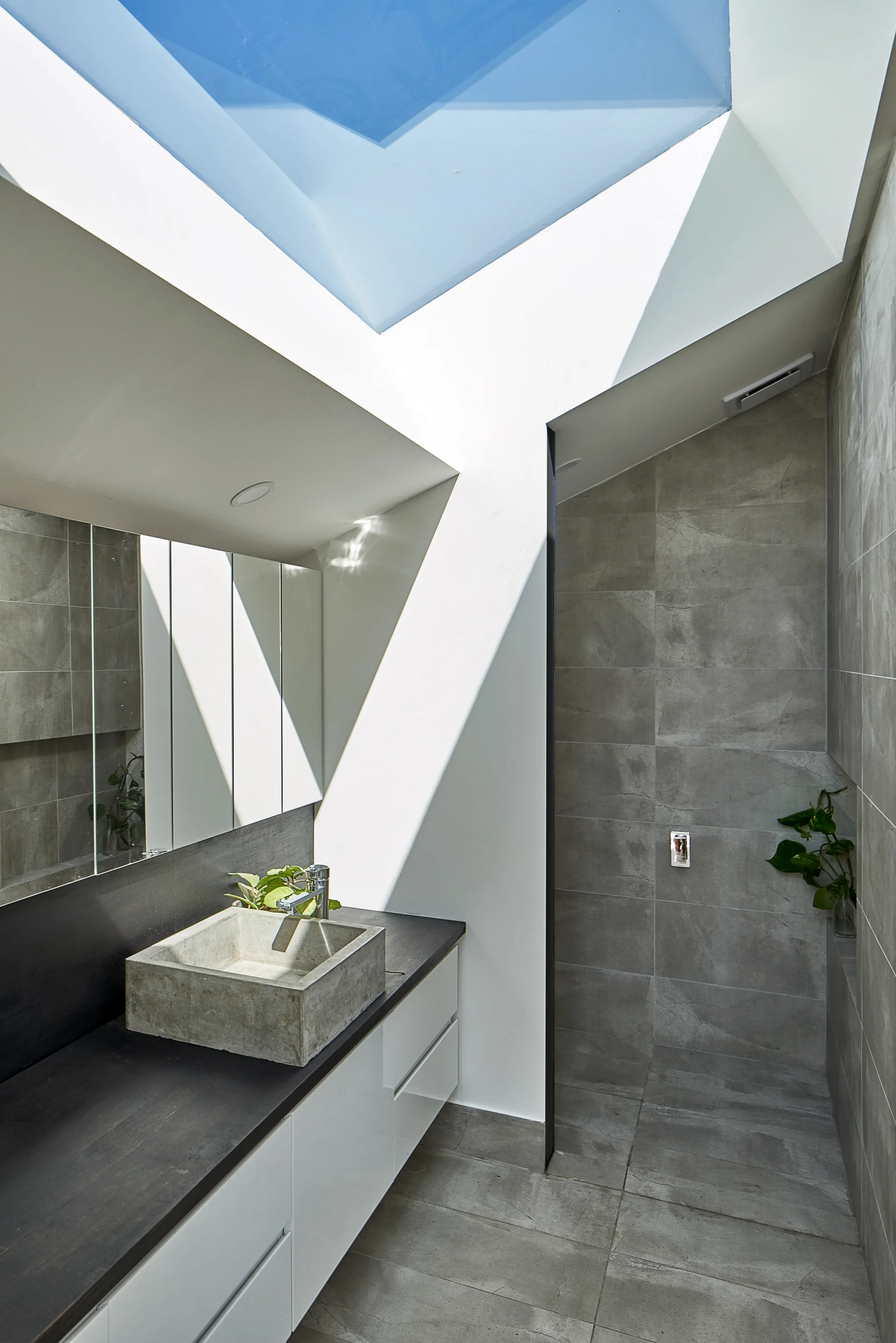 A modern gray tiled bathroom featuring a concrete vessel sink on a dark countertop beneath a bright angled skylight.