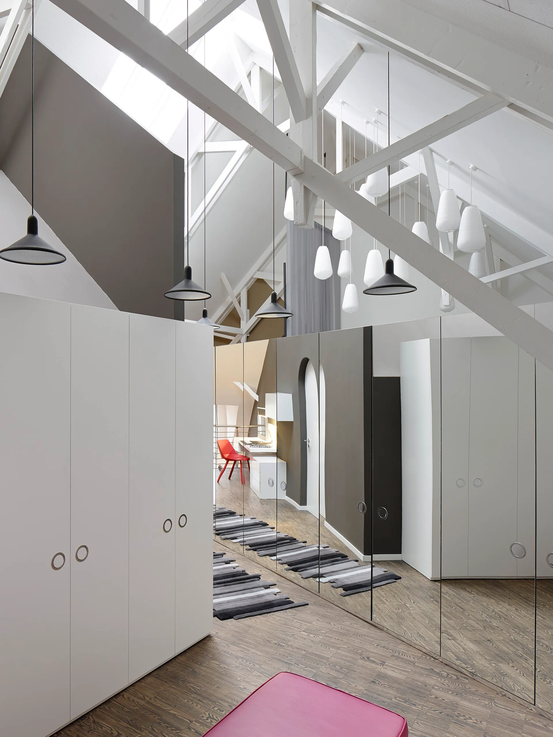 A modern attic bedroom featuring white wardrobes, exposed ceiling beams, and a large mirrored wall reflecting a study desk and striped rug.