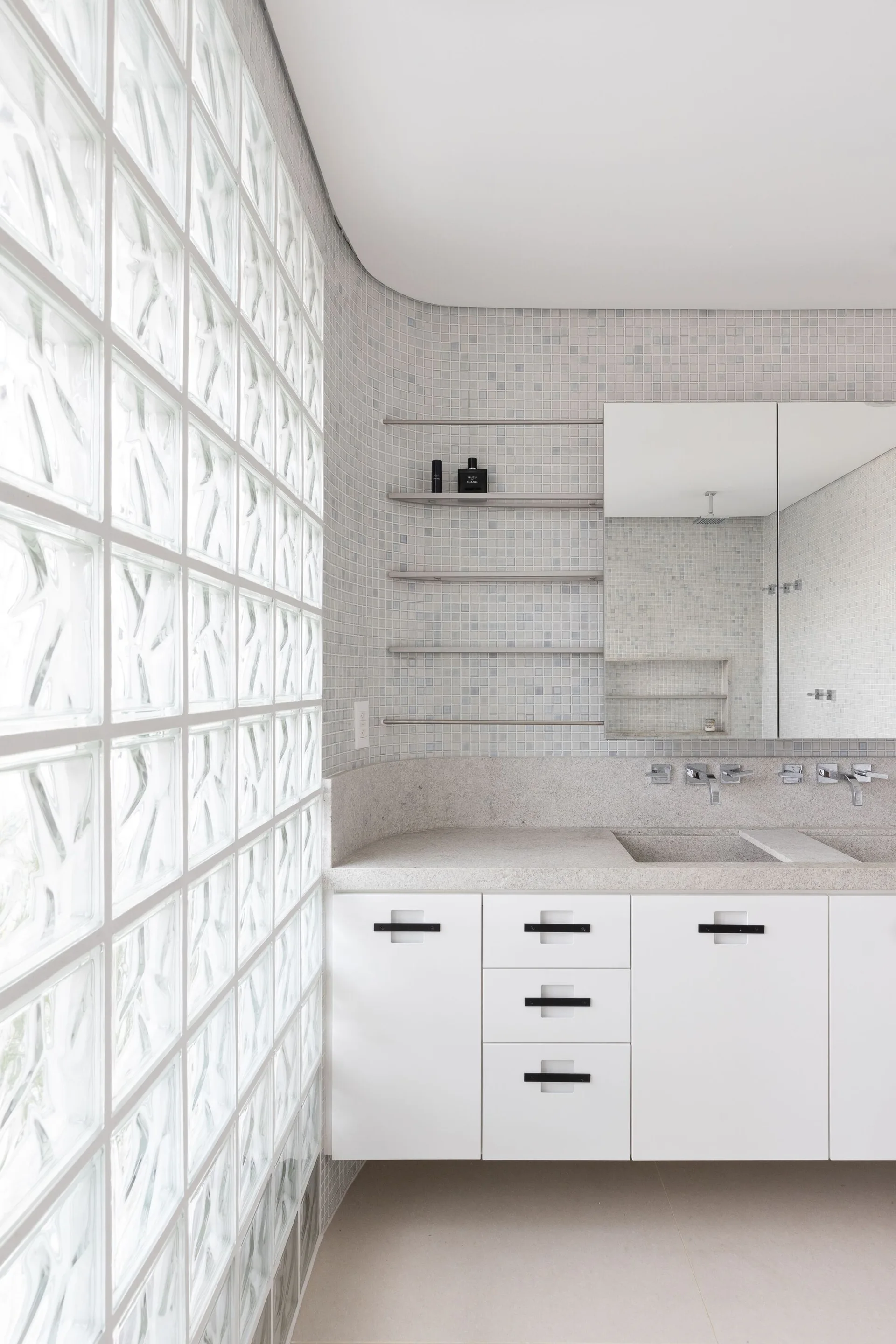 A white bathroom vanity with a light stone countertop next to a translucent glass block wall and a small mosaic tiled backsplash.
