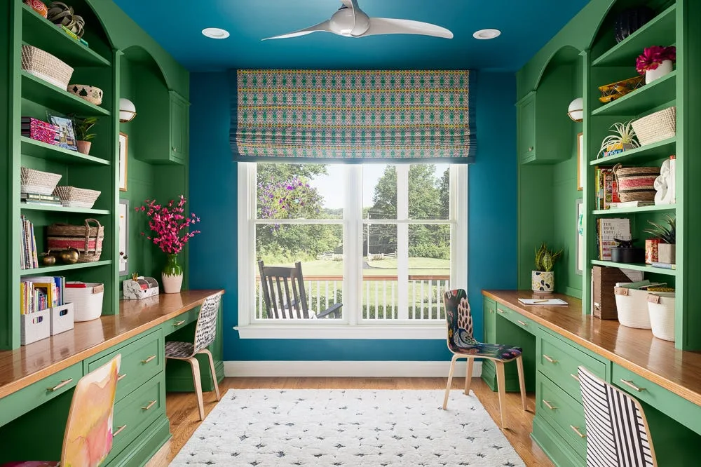 A symmetrical home office featuring green built-in desks and shelving, wood countertops, blue walls, a center window, and two patterned chairs.