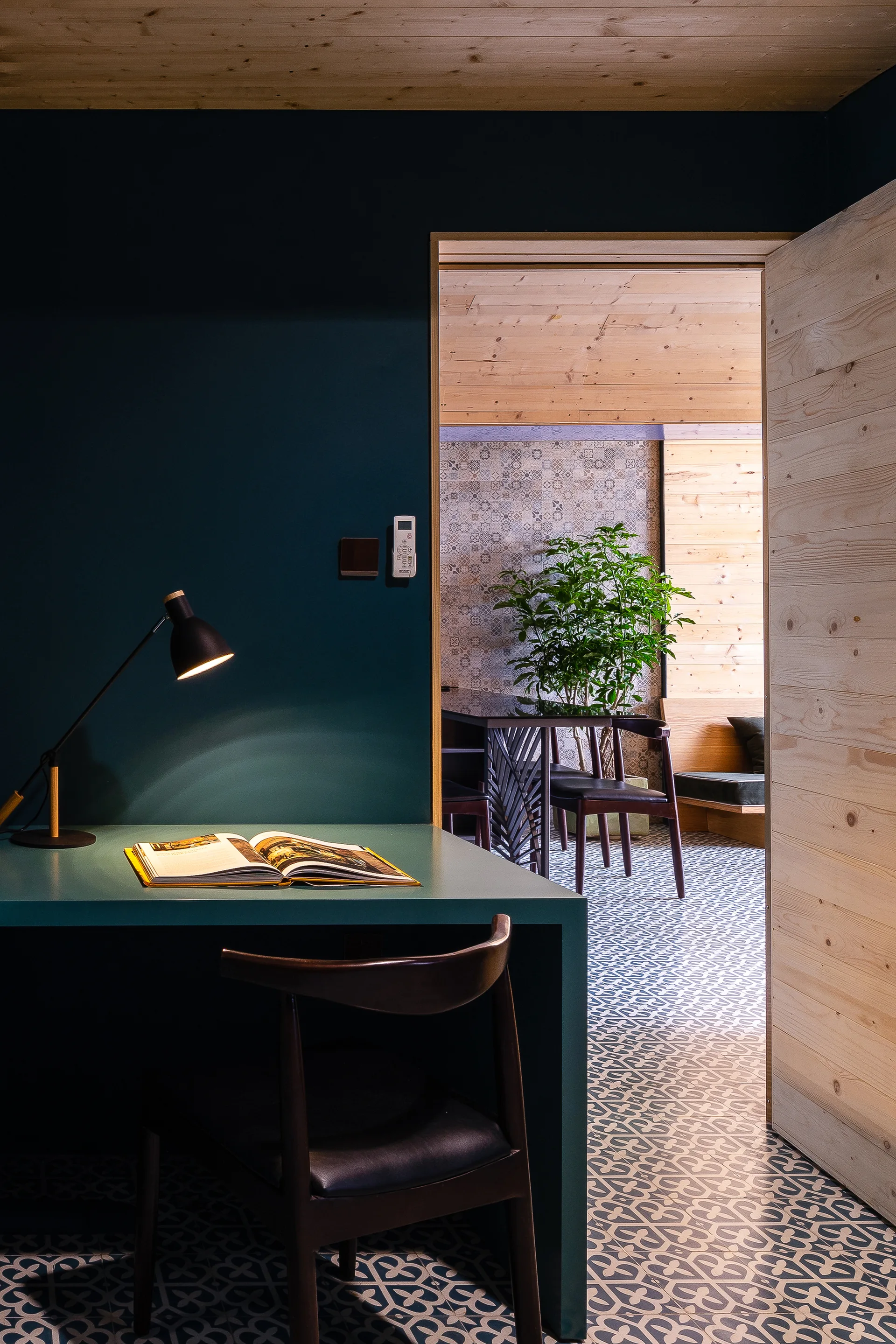 A teal desk and chair sit against a dark teal wall beneath a light wood ceiling with patterned floor tiles continuing into an adjacent room.