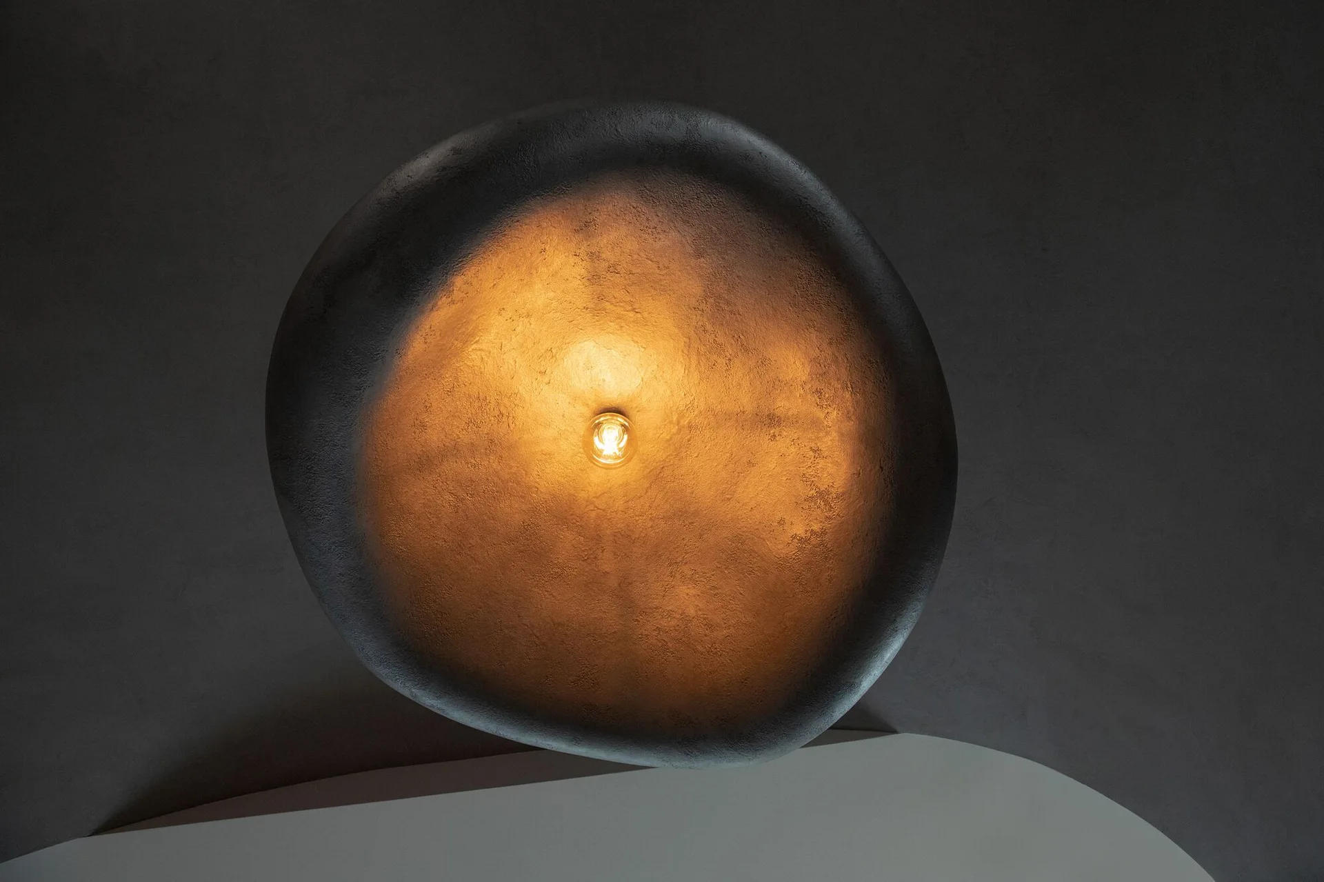 A low-angle view of a textured, bowl-shaped pendant light glowing with a central bulb against a dark grey ceiling.