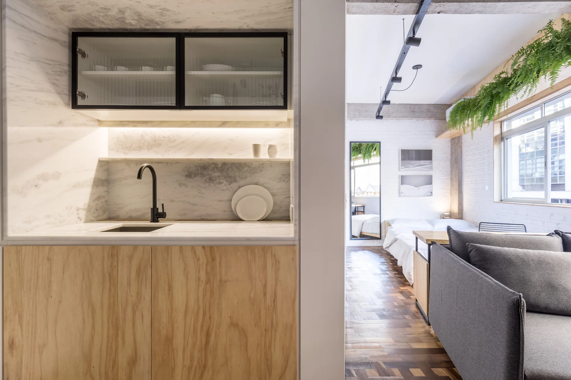 A modern studio apartment features a marble kitchenette with light wood cabinets, a grey sofa, and block-pattern parquet flooring.