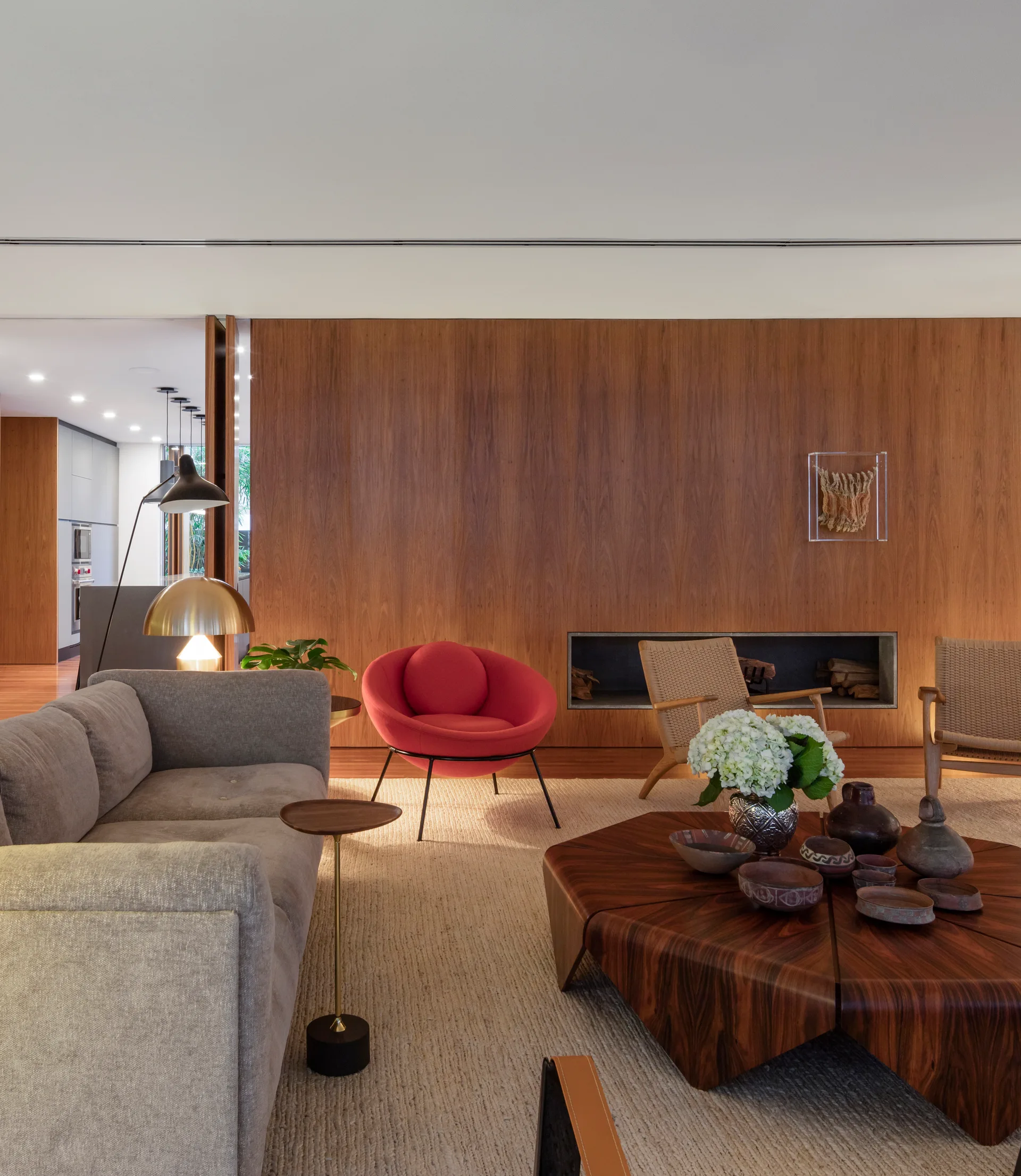 A modern living room featuring a large wood-paneled wall, a gray sofa, a red round chair, and a low sculptural wood coffee table on a textured rug.