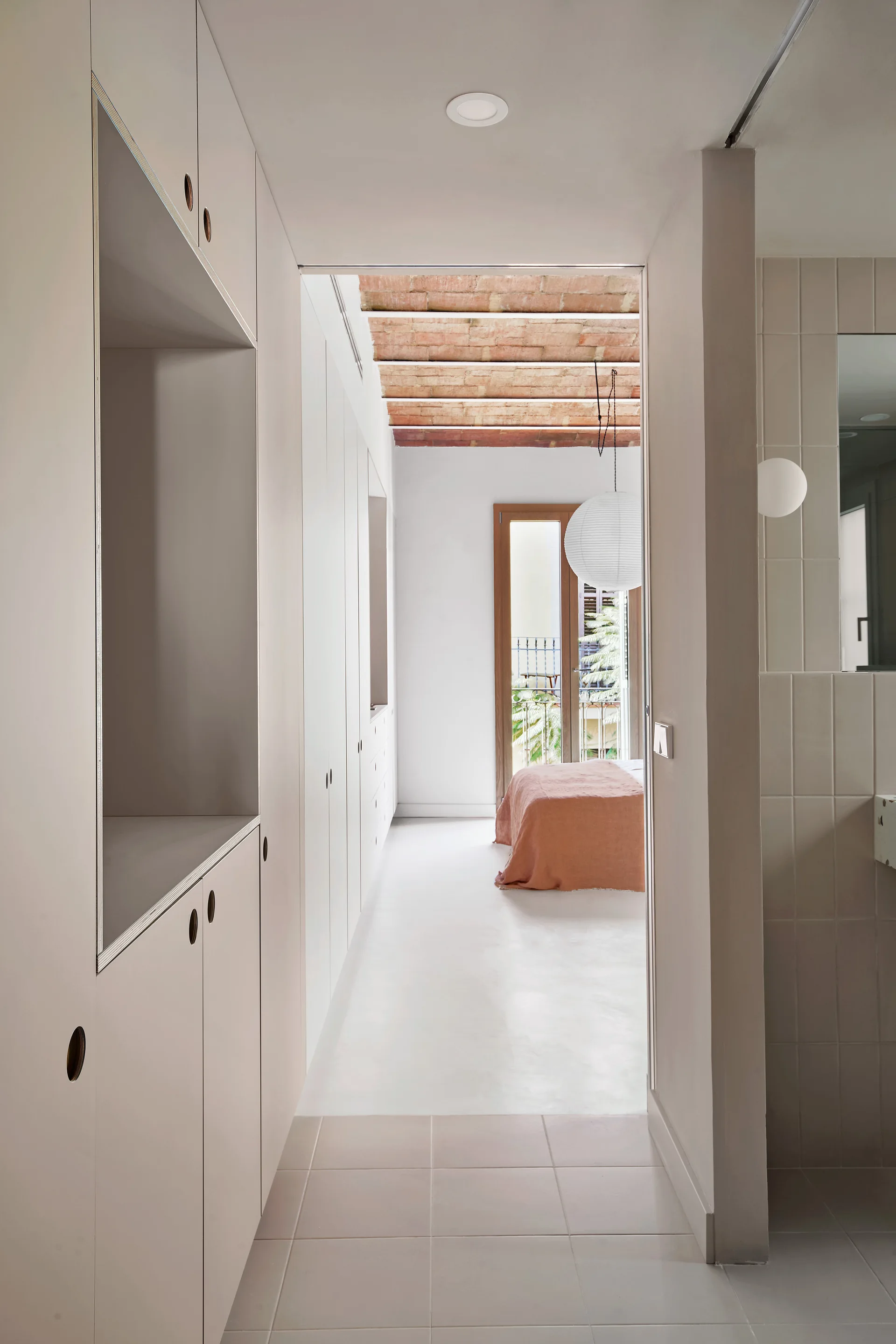 A bright hallway with custom built-in cabinets leads into a bedroom featuring an exposed brick vaulted ceiling and a bed with terracotta linen.