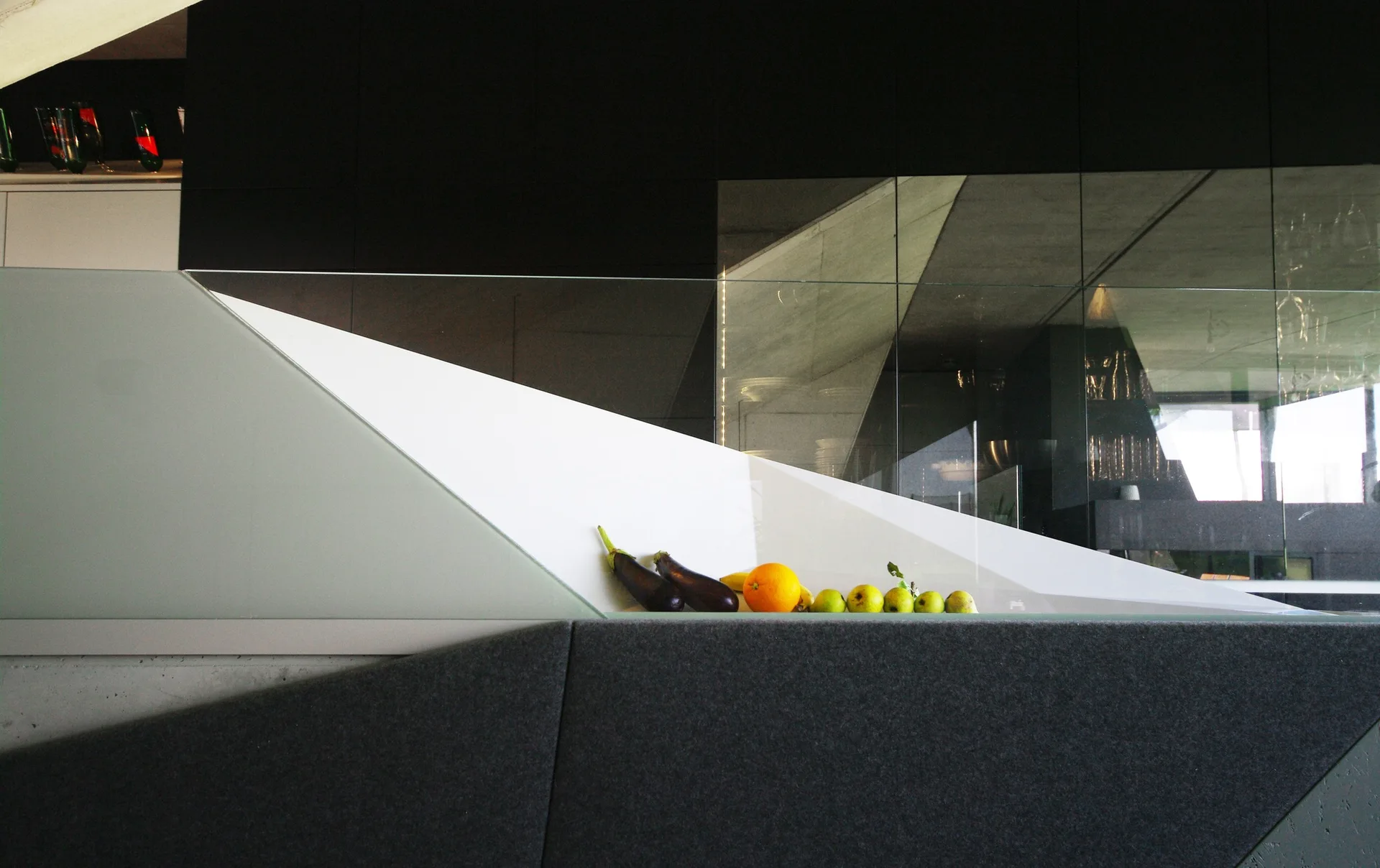 A minimalist interior stairwell featuring a white diagonal railing with glass panels, dark grey fabric walls, and fresh fruit resting on a ledge.