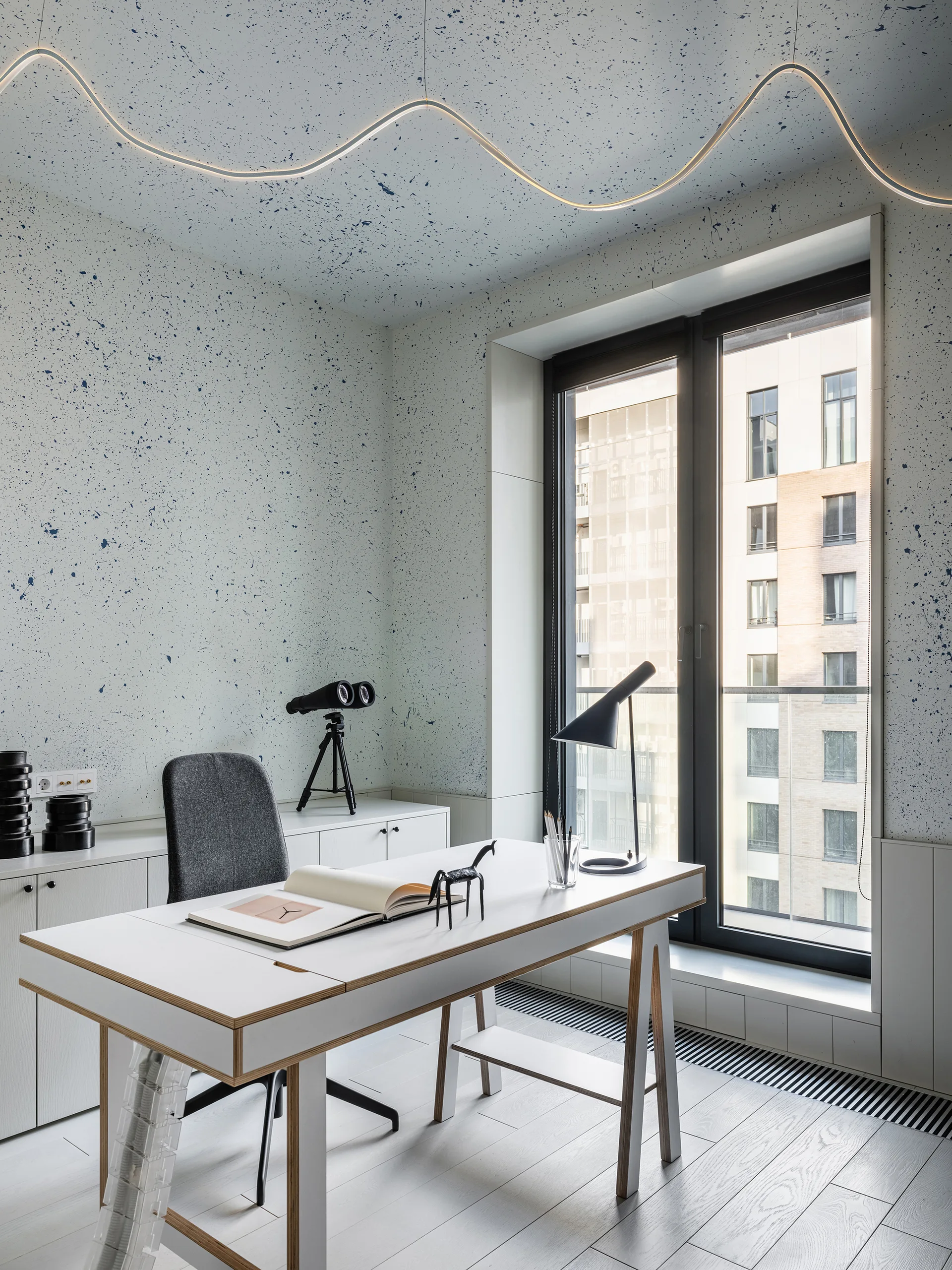 A modern home office features a white writing desk, a grey chair, and walls and ceiling covered in a blue paint splatter pattern.