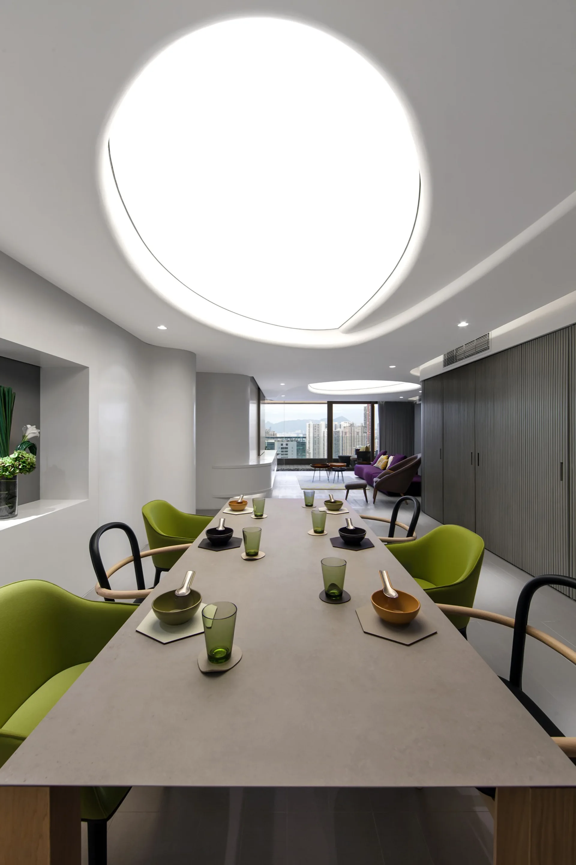 A long grey dining table set with dishes and green glasses is surrounded by lime green chairs beneath a massive circular recessed ceiling light.