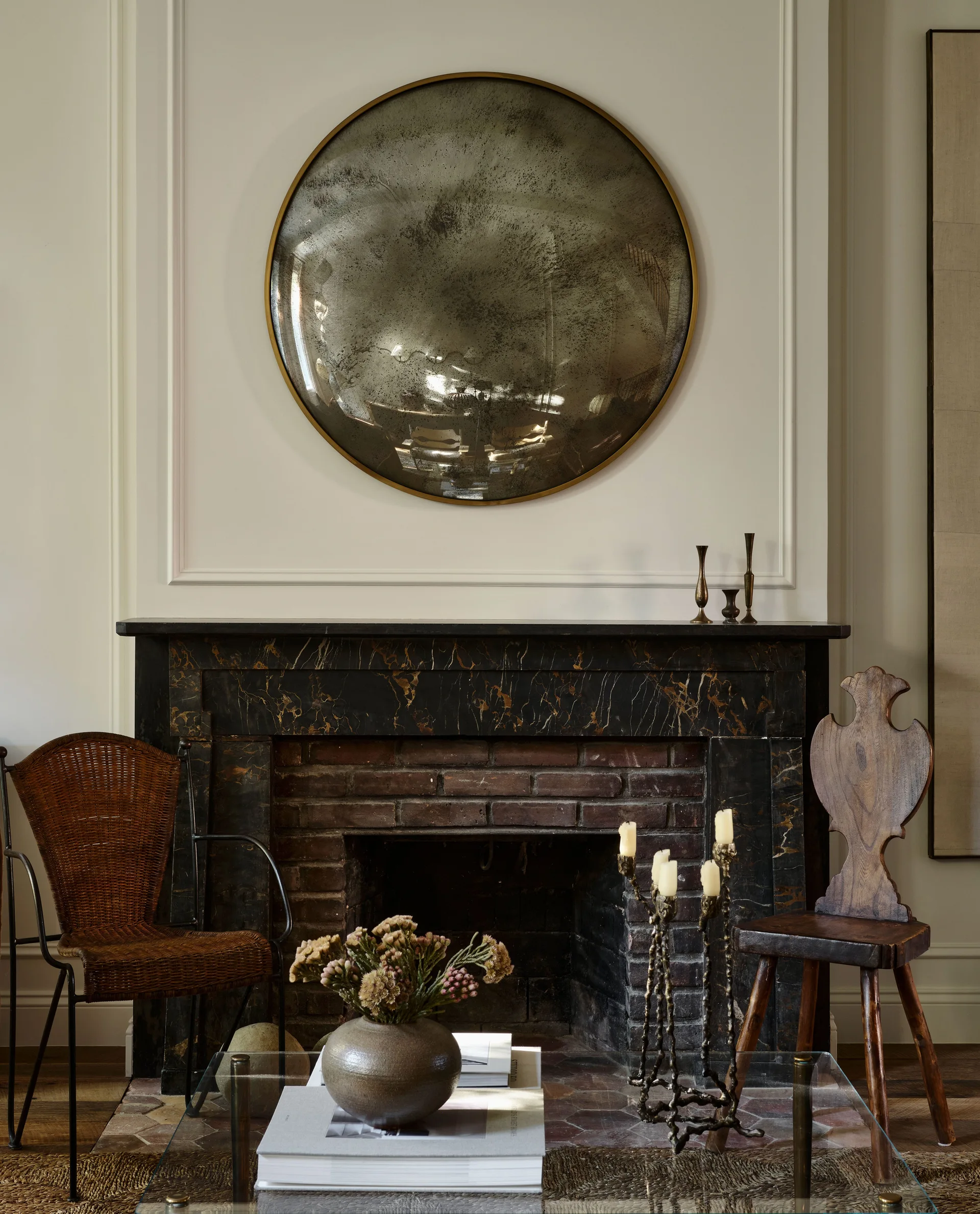 A dark marble fireplace with a brick interior features a large round aged mirror on the wall above, flanked by two rustic wooden chairs.