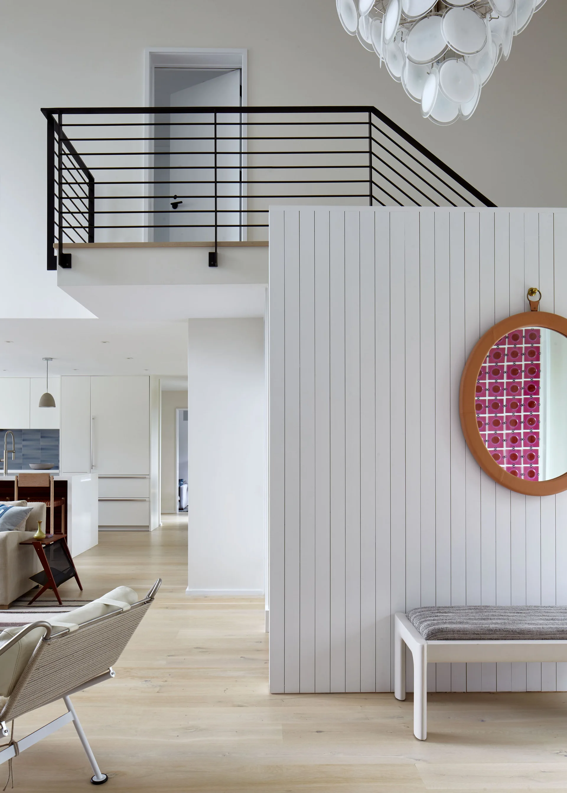 A white entryway features a vertically slatted accent wall with a round orange-rimmed mirror, a small bench, and a black staircase railing above.
