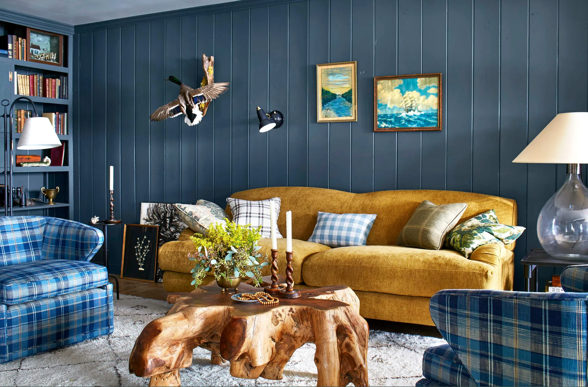 A living room features a mustard yellow sofa and two blue plaid armchairs set against a dark blue paneled wall with a live edge wood coffee table.